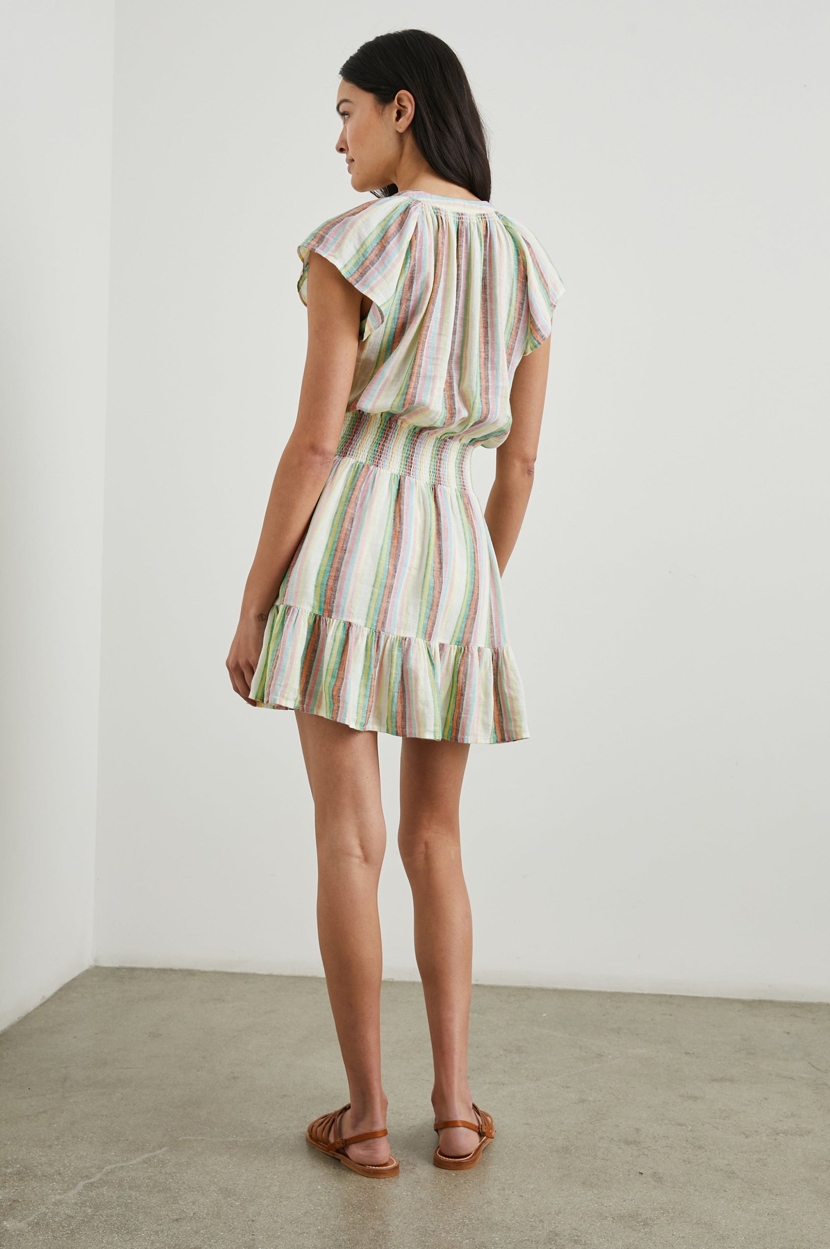Augustine Short Sleeve Mini Dress with Flutter Cap Sleeves, a V Neck, a Smocked Waist, a Ruffled Hem, and a Placket with Buttons in Palais Stripe Colorway, Vertical Stripes in Green, Yellow, Orange, Brown, Pink, Blue and white - Back Full Body View Featured on Model