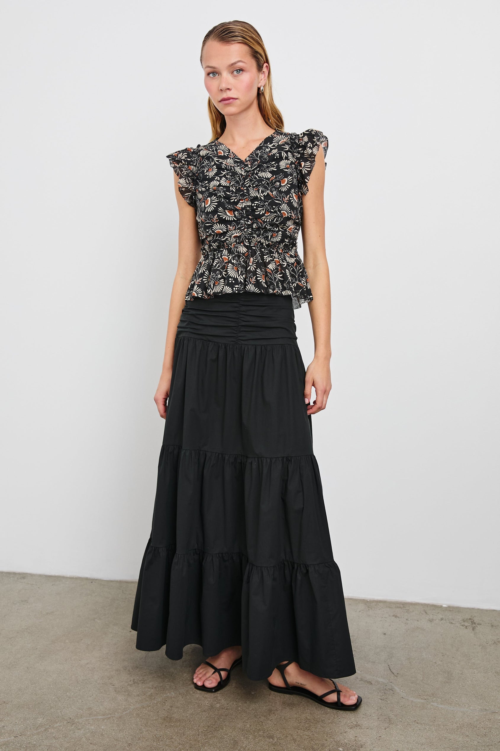 Cali Top with Flutter Sleeves, a V Neckline, Horizontal pleats along the bodice and vertical pleats at the hem in Cassia Colorway, with white and orange flowers on black - Front Full Body View Featured on Model