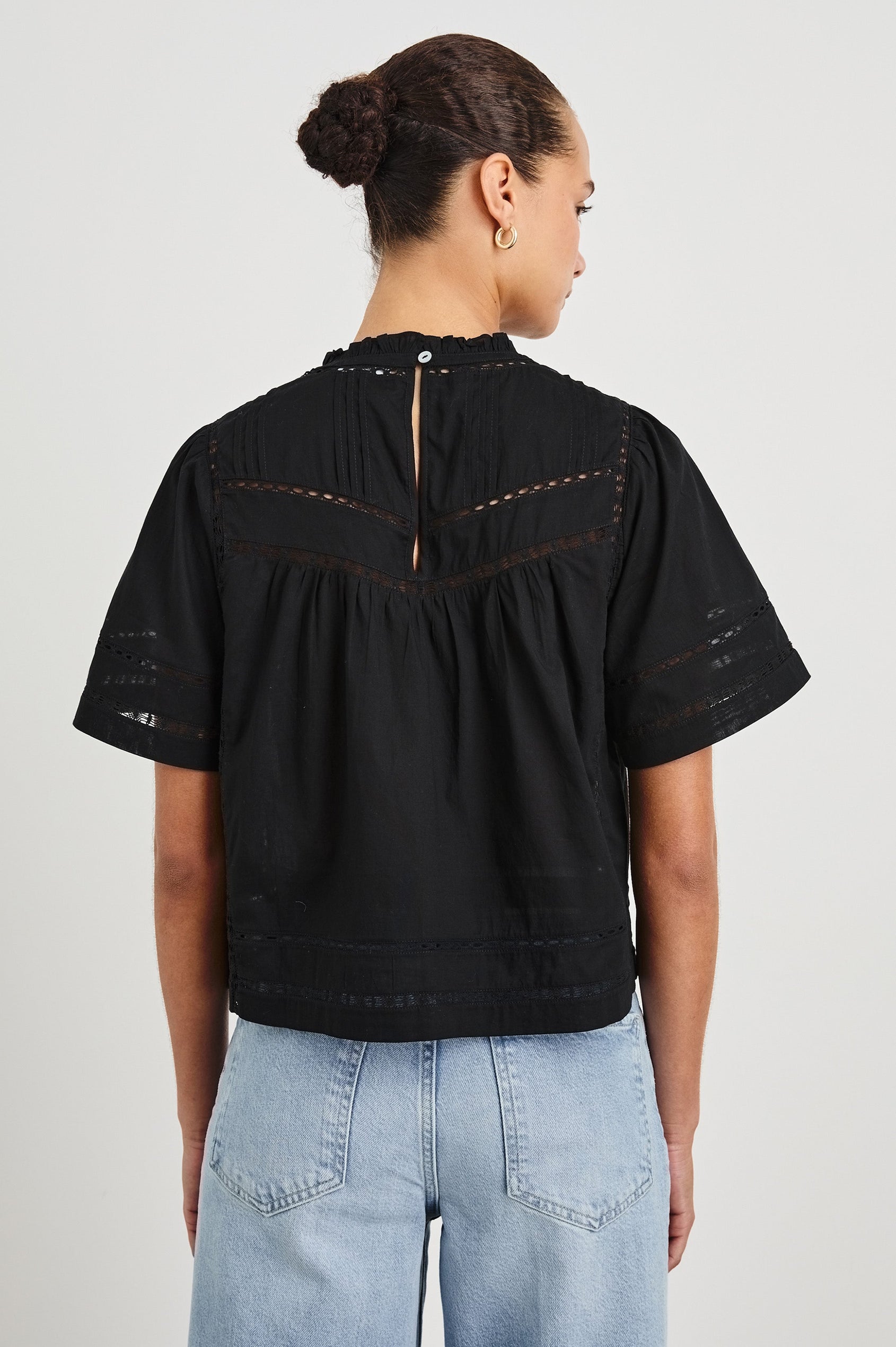 Back view of female model wearing our Daisy Top in black. This is a crewneck baby doll blouse, with ruffles at the collar. Styled with light wash denim and a simple belt.