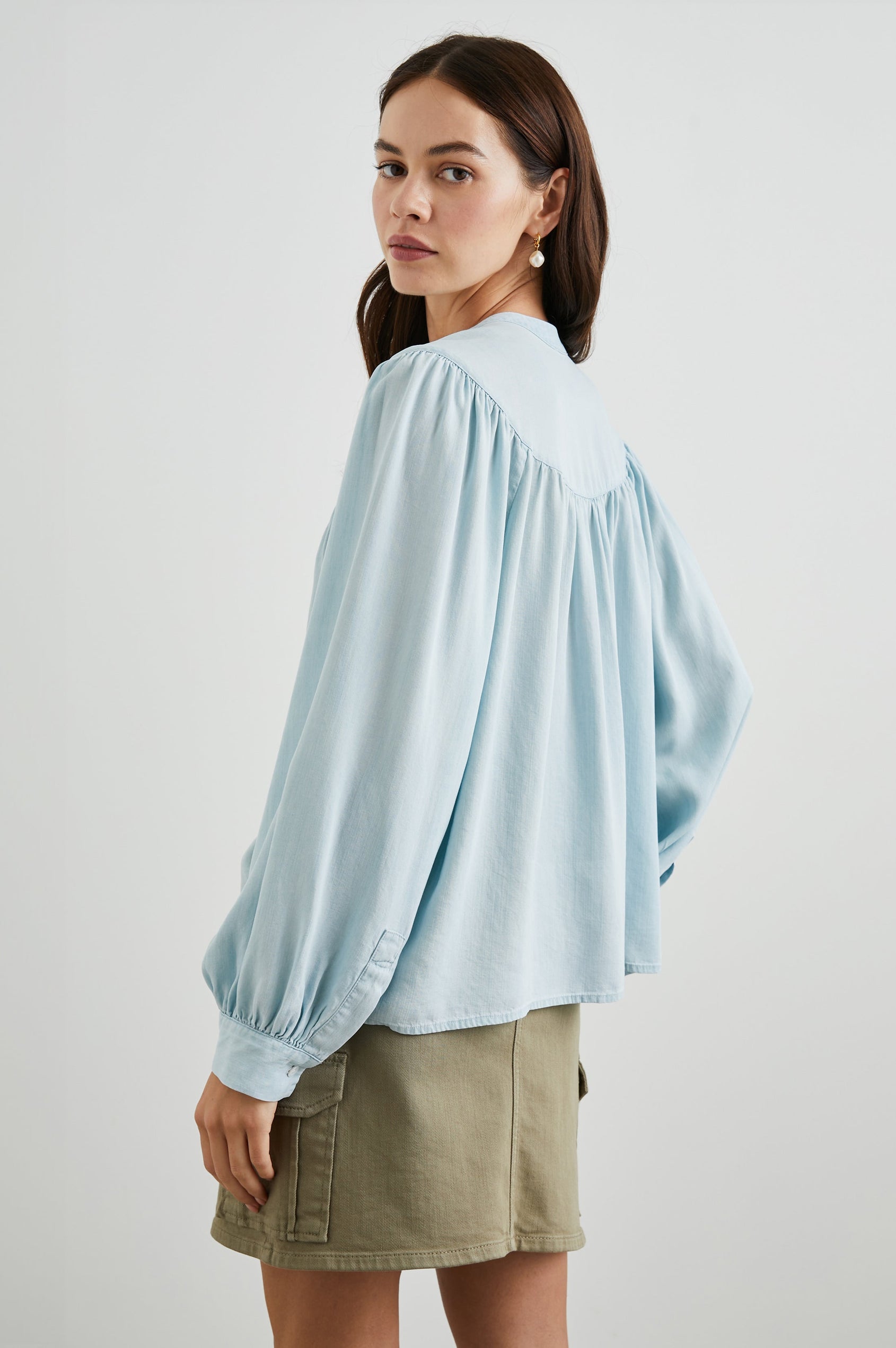 Fable Long Sleeve Top with a V Neck with a Placket, Buttoned Cuffs, and Pleating Down From the Yoke in Light Vintage Colorway, a Blue - Back View Featured on Model