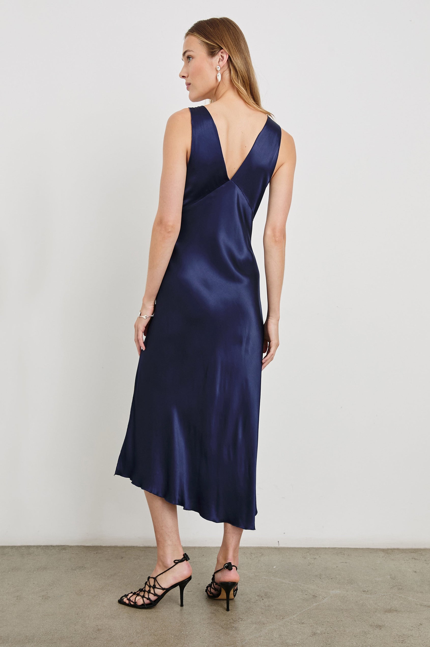 Monique Sleeveless Midi Dress with an Asymmetrical Hem Line, Side Slit, Deep V Neck, and Wide Shoulder Straps in Navy Colorway - Back Full Body View Featured on Model