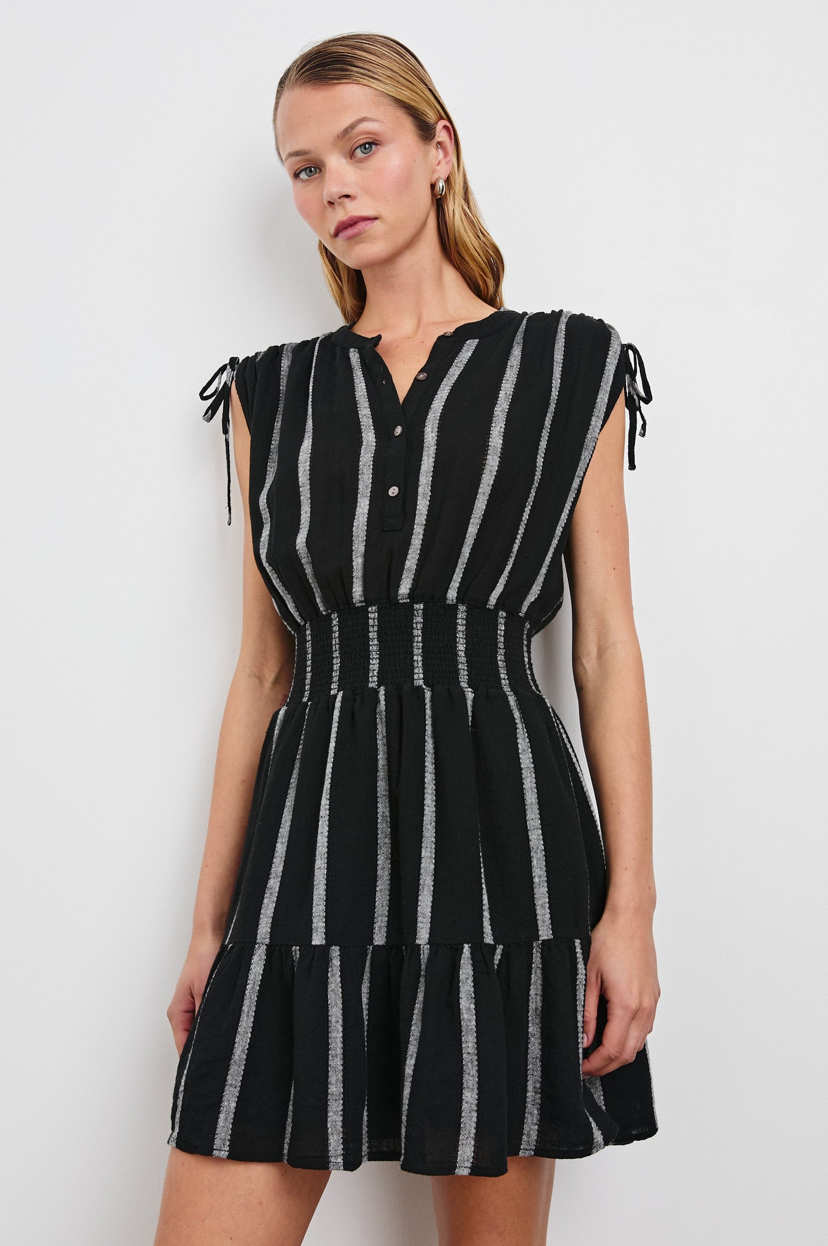 Samina Above-Knee Sleeveless Dress with drawstrings at the Arm Openings, Buttons along the Bodice, A Cinched Waist and a Tiered Skirt in Ash Leno Stripe Colorway, with Grey vertical stripes on black - Front View Featured on Model