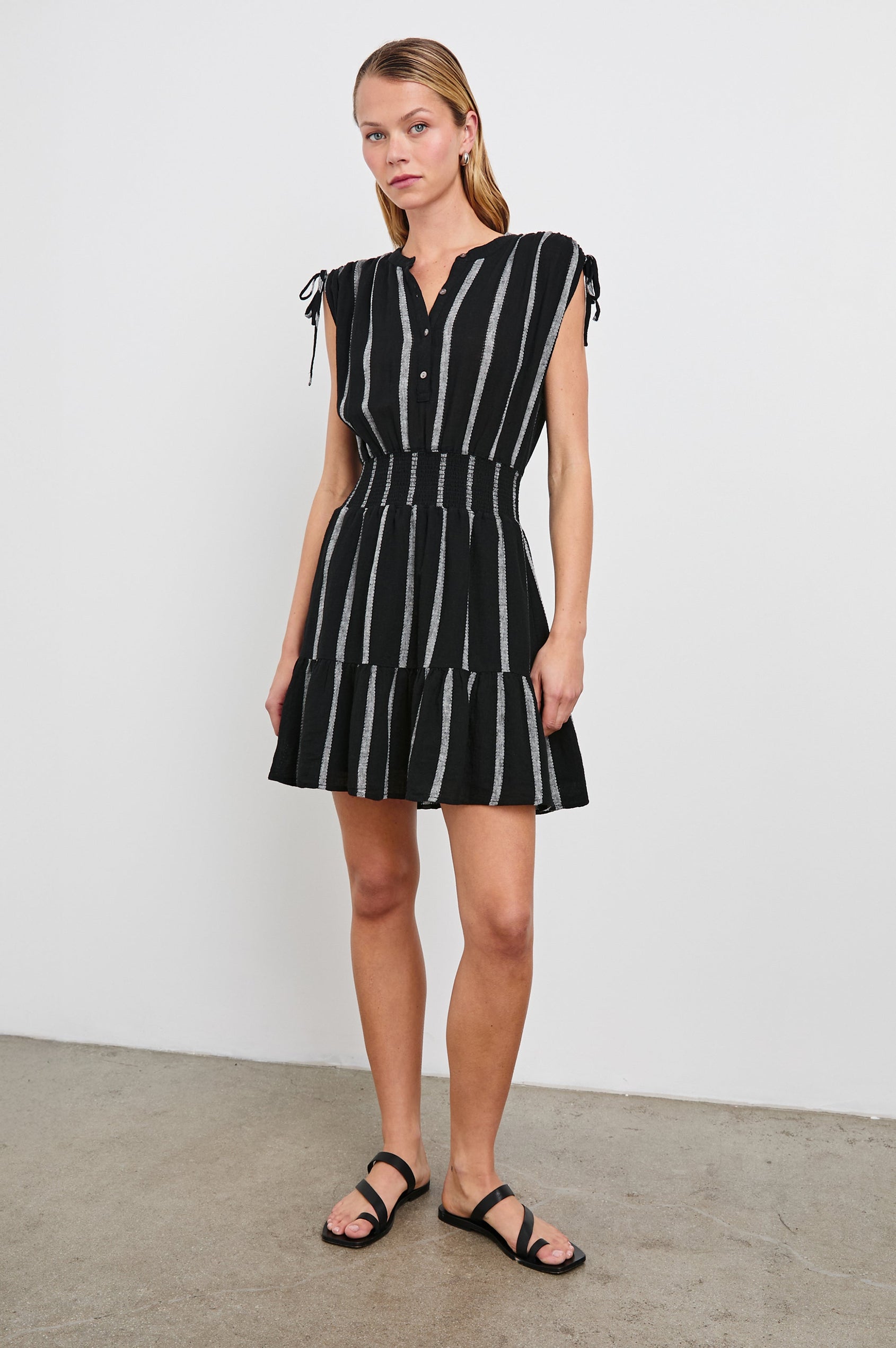 Samina Above-Knee Sleeveless Dress with drawstrings at the Arm Openings, Buttons along the Bodice, A Cinched Waist and a Tiered Skirt in Ash Leno Stripe Colorway, with Grey vertical stripes on black - Front Full Body View Featured on Model