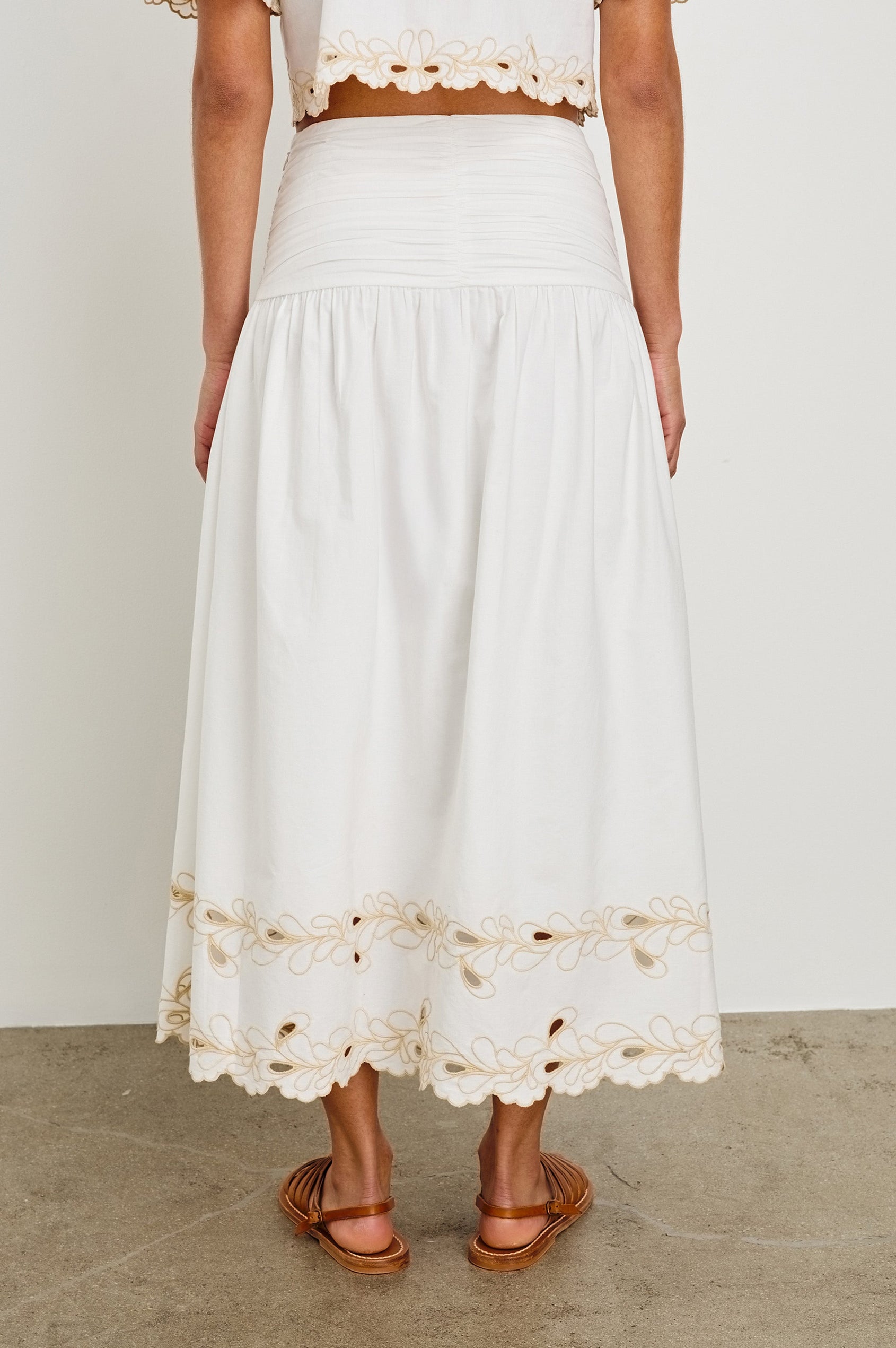 Back image of model wearing a white, cotton poplin midi skirt with a lightly gathered waist that cinches and drops at the hips. Details include beige embroidered eyelets at the hem. Worn with brown sandals.