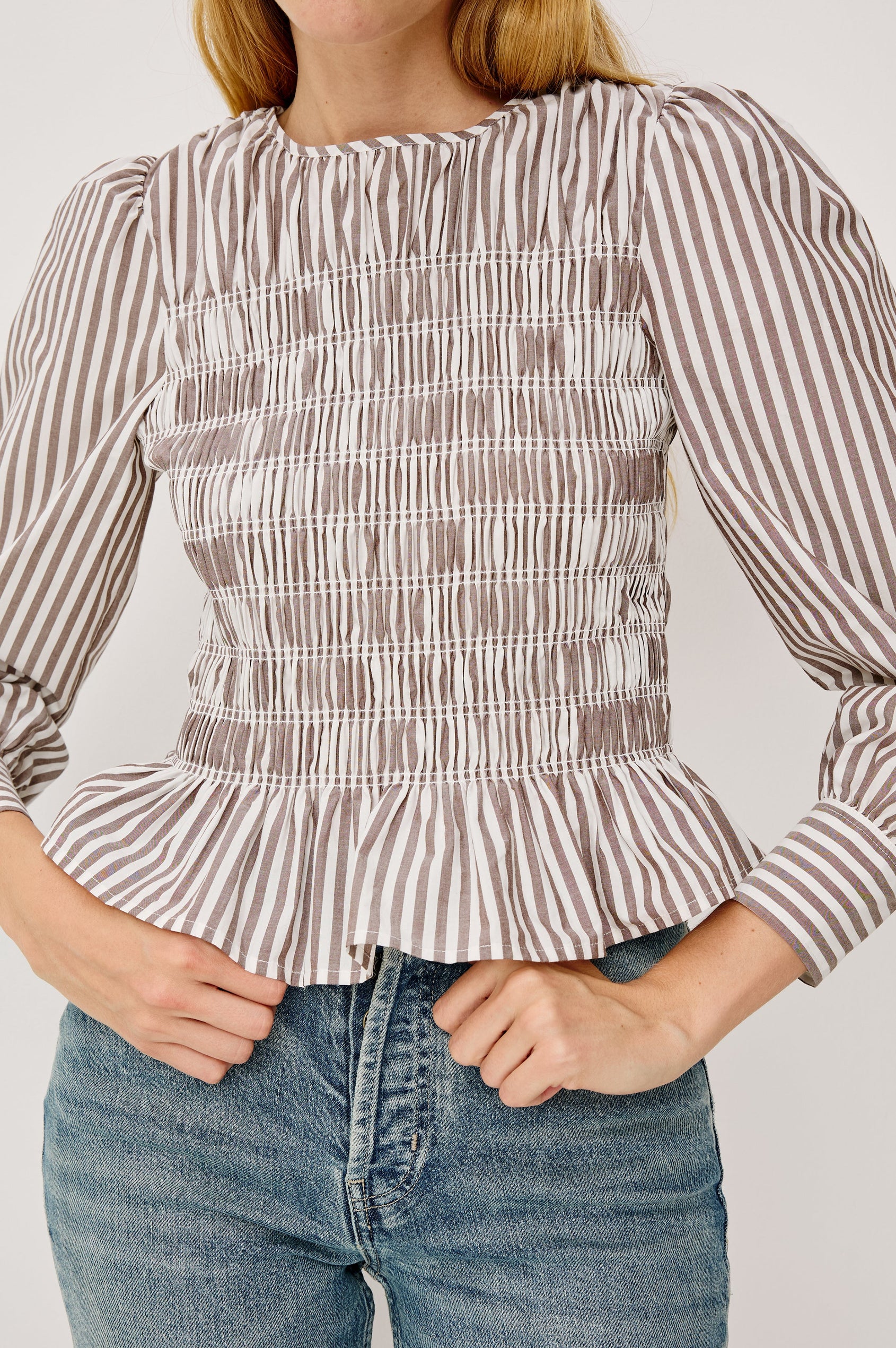 Tabi is a long sleeve crew neck top with puff sleeves and buttons at cuff, the bodice is pleated with a peplum hem in an Espresso Ivory Stripe colorway. Close Up view featured on model.