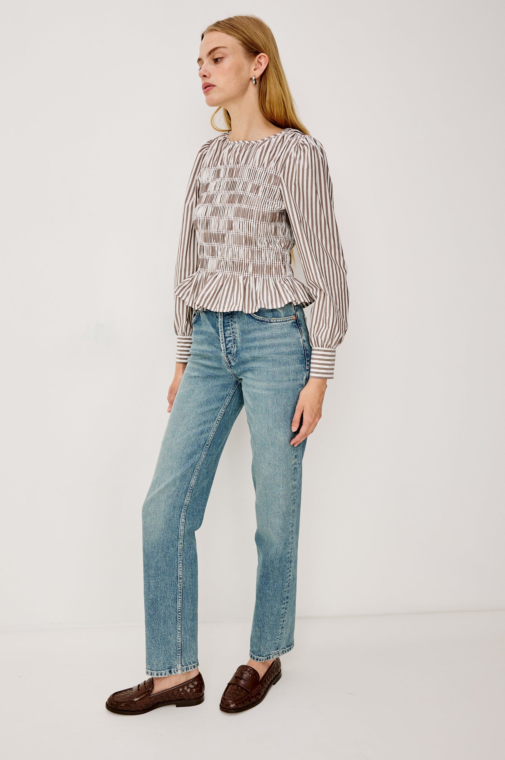 Tabi is a long sleeve crew neck top with puff sleeves and buttons at cuff, the bodice is pleated with a peplum hem in an Espresso Ivory Stripe colorway. Full Body Front view featured on model.