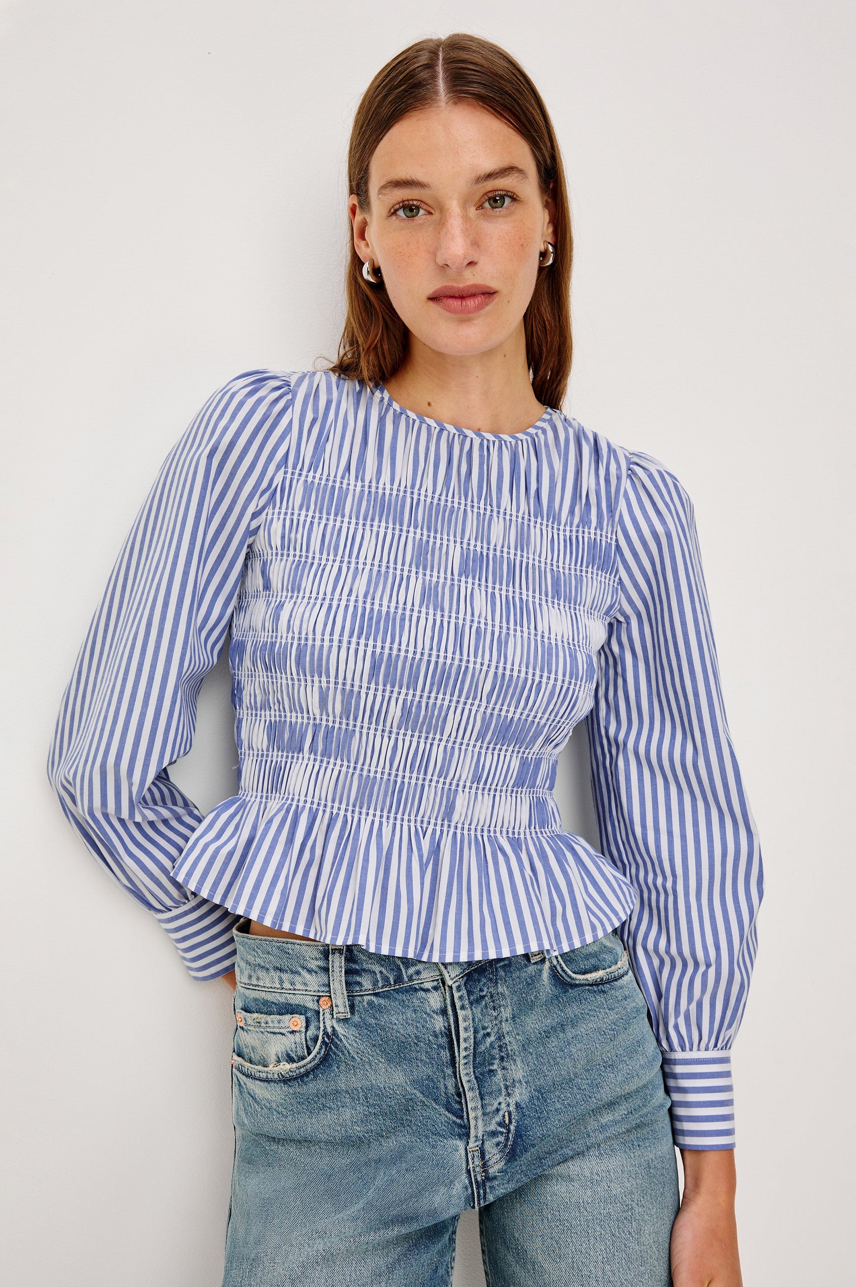 Tabi is a long sleeve crew neck top with puff sleeves and buttons at cuff, the bodice is pleated with a peplum hem in a blue Mariner White Stripe colorway. Front view featured on model.