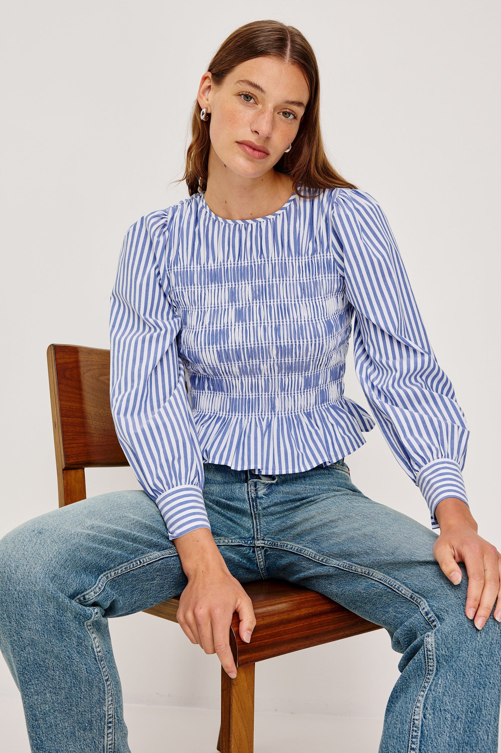 Tabi is a long sleeve crew neck top with puff sleeves and buttons at cuff, the bodice is pleated with a peplum hem in a blue Mariner White Stripe colorway. Front seated view featured on model.