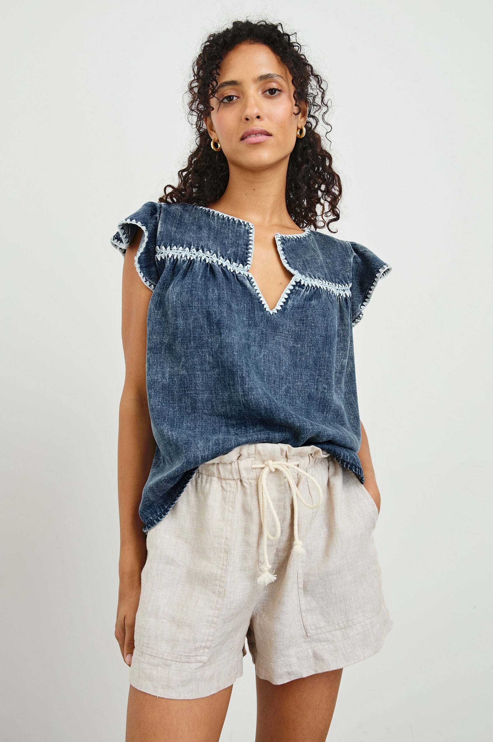 Tafia Top with an Open V-neck, Short Ruffle Cap Sleeves, and a White Blanket Stich along the Neckline, Cuffs and Yoke Seam in Classic Blue Colorway - Front Half-Tucked View Featured on Model