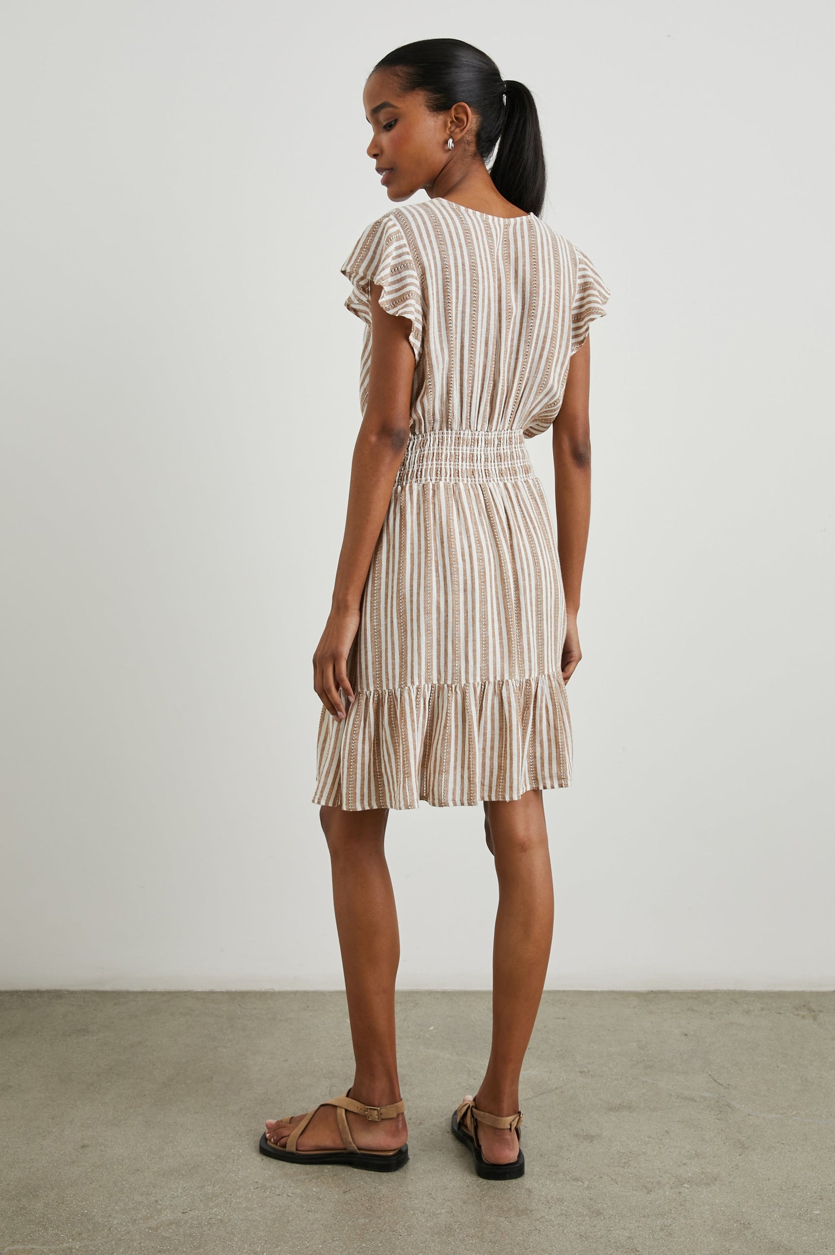 Tara Short Sleeve, Above the Knee Dress with a Smocked Waist, V Neck, Flutter Cap Sleeves, and Tiered Skirt with a Ruffled Hem in Palo Santo Stripe Colorway, with Textured Brown Vertical Stripes on White - Back Full Body View Featured on Model