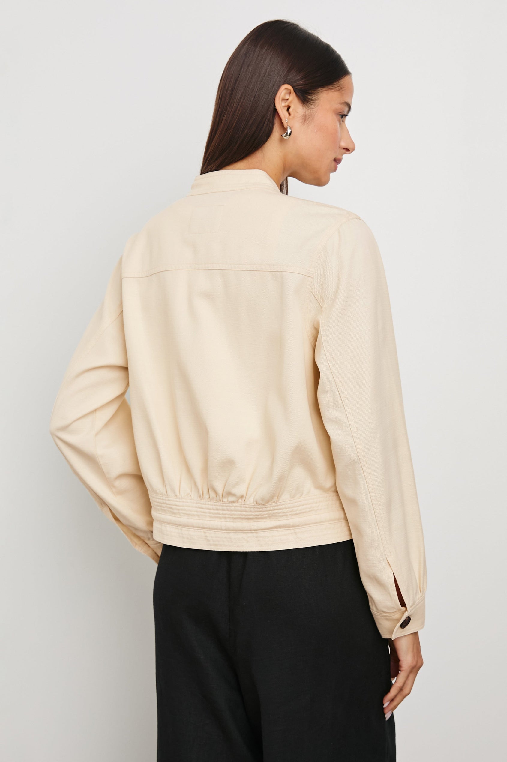 Alma Jacket with Button Closure, Mandarin Collar, Two Large Chest Button Patch Pockets and a Drawcord at the Hem in Ecru Colorway, with Dark Buttons - Back View Featured on Model