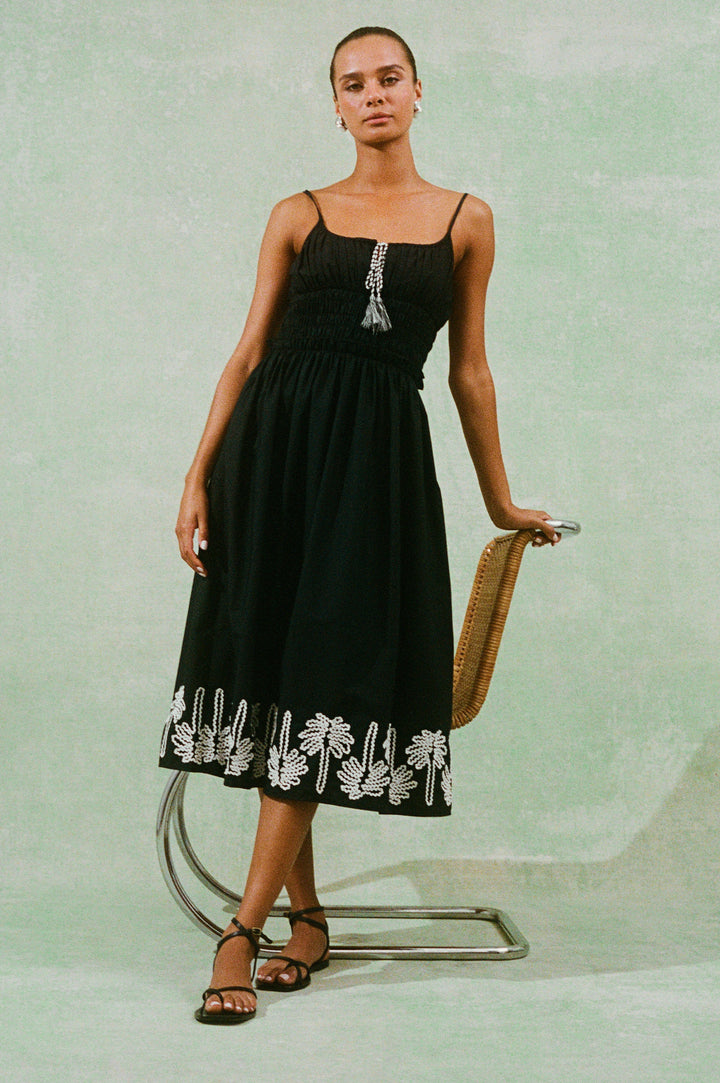 Baja Black Dress with Ivory Palm Tree Stitching at the skirt hem and neckline cord ties - front view on model with chair prop