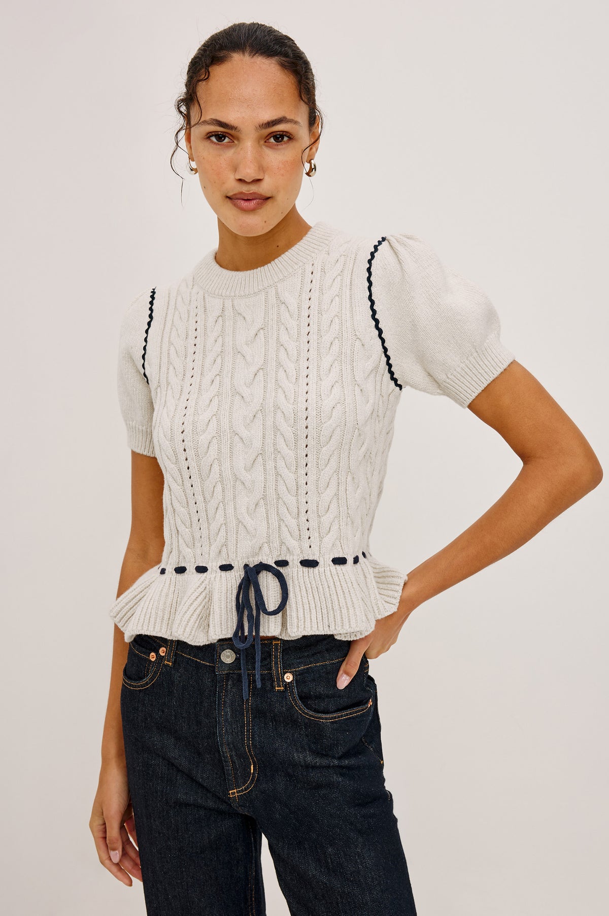 Chandi Sweater in Cable Knit With Short Puff Sleeves, Fitted SIlhouette, Soft Flared Peplum Hem, and Contrast Serpentine Trim and Interlocking Drawstring Ties in Heather Oat Colorway - Full Body Front View Featured on Model