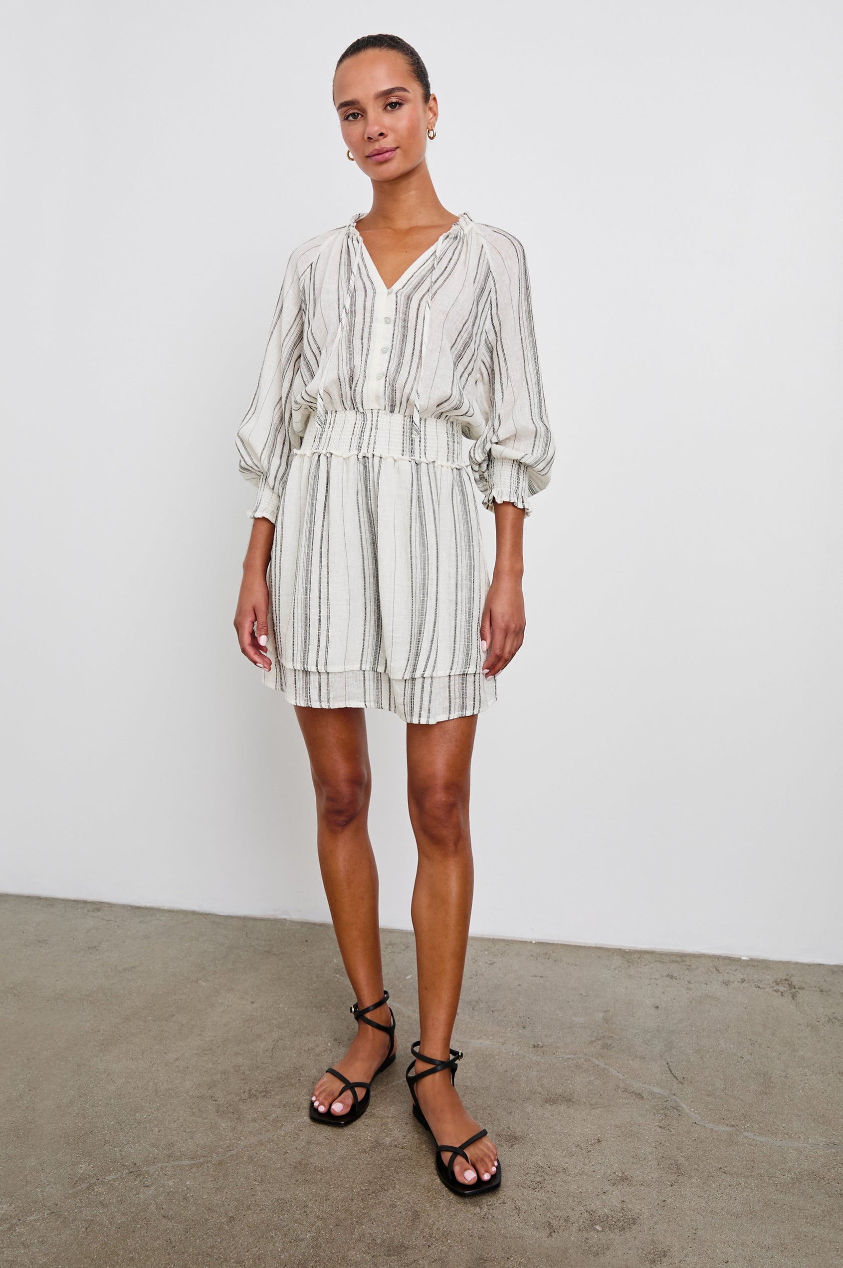 Crystal Mini Dress with Mid-Length Sleeves, Shirred Cuffs and Waist, a V Neck, Ties at the Collar, and Buttons down the Bodice in Basque Stripe Colorway, Thin Black Vertical Stripes on White - Front Full Body View Featured on Model 