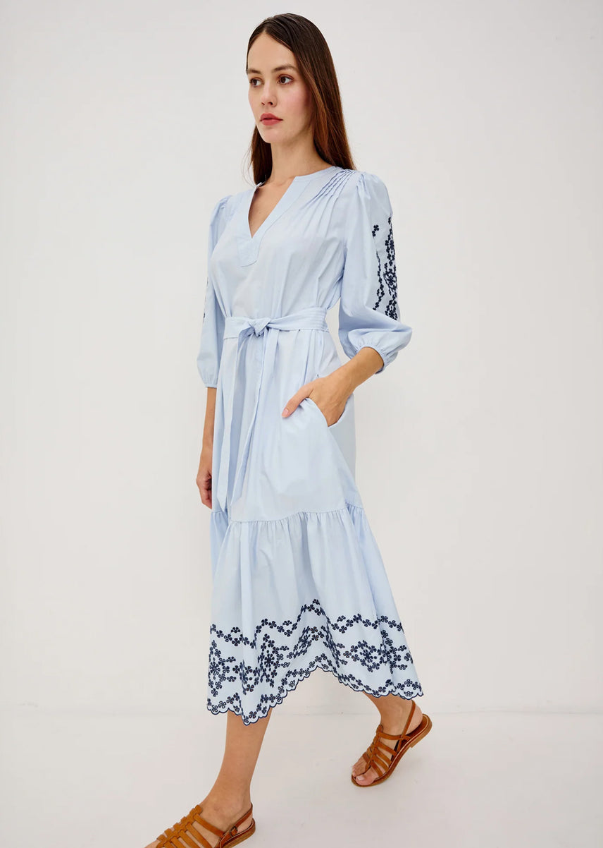 A woman modeling a light blue midi dress with dark blue floral embroidery on the sleeves and hem, paired with brown leather sandals, against a plain white studio background.