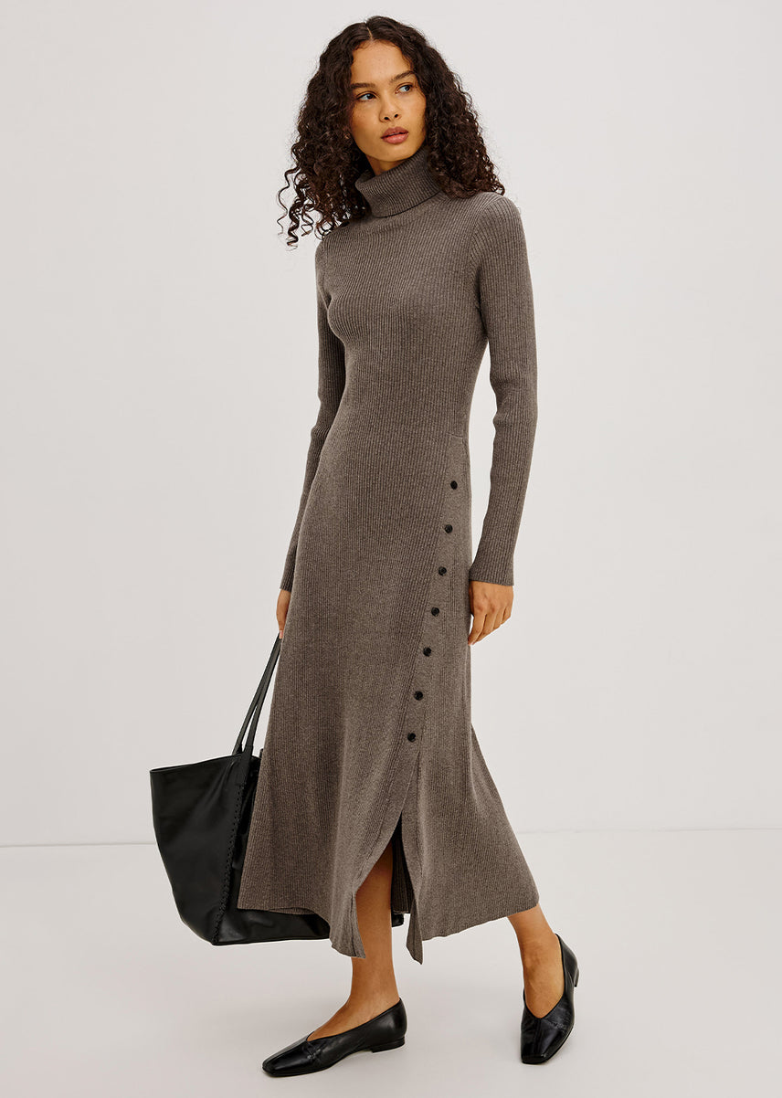 A woman modeling a taupe brown ribbed knit mock-neck midi dress with a side button placket, styled with a black tote bag and black leather loafers against a light gray studio background.