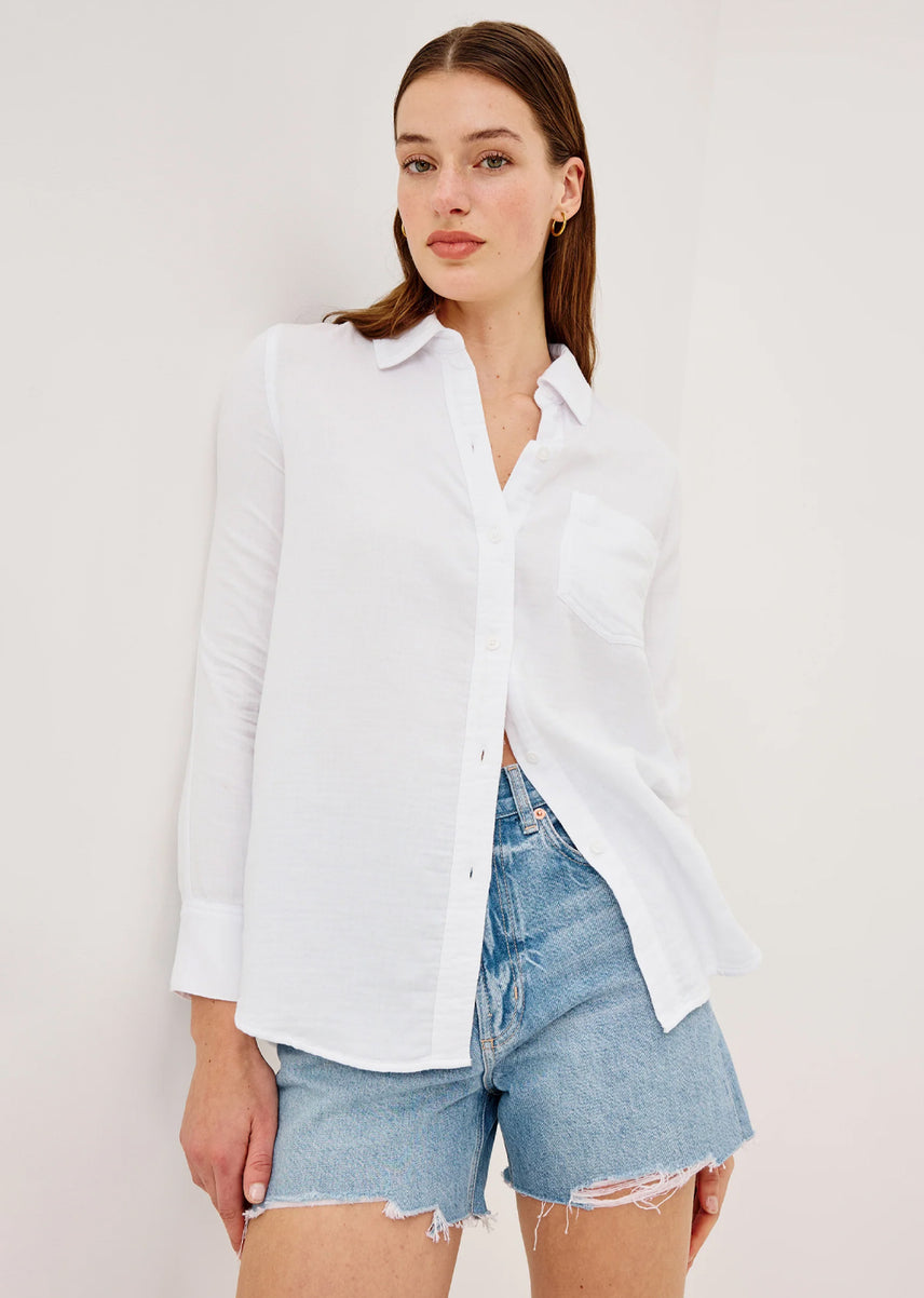 A woman modeling a white long-sleeve button-up shirt with a chest pocket, paired with light-wash denim shorts, against a plain off-white background.