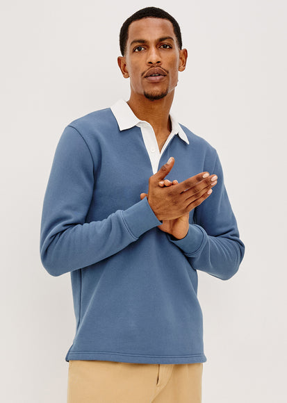 A man wearing a dusty blue long-sleeve polo shirt with a white collar, paired with beige trousers, standing against a light gray studio background.