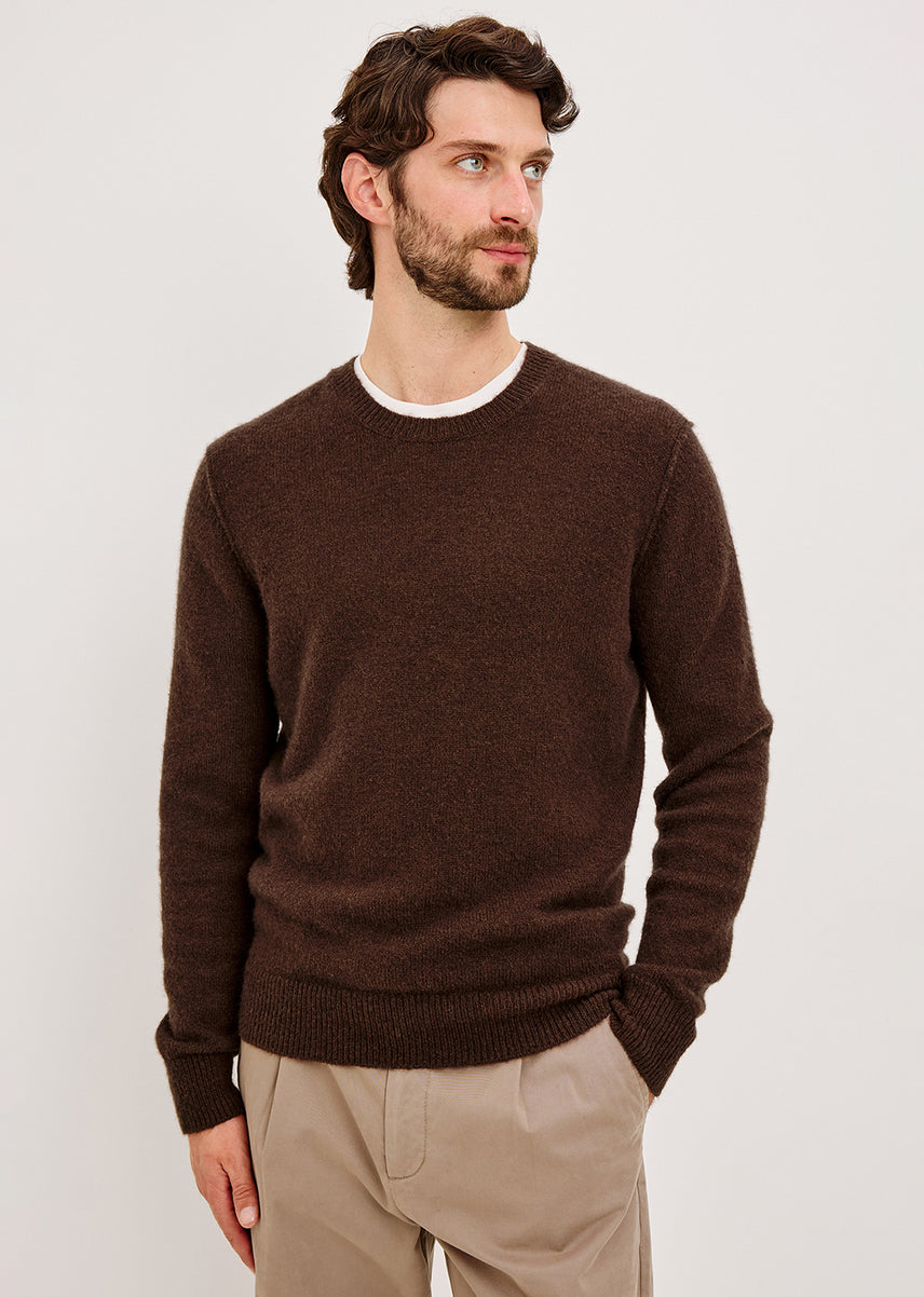A man wearing a dark brown crew-neck knit sweater over a white undershirt and beige trousers, standing against a light gray studio background.