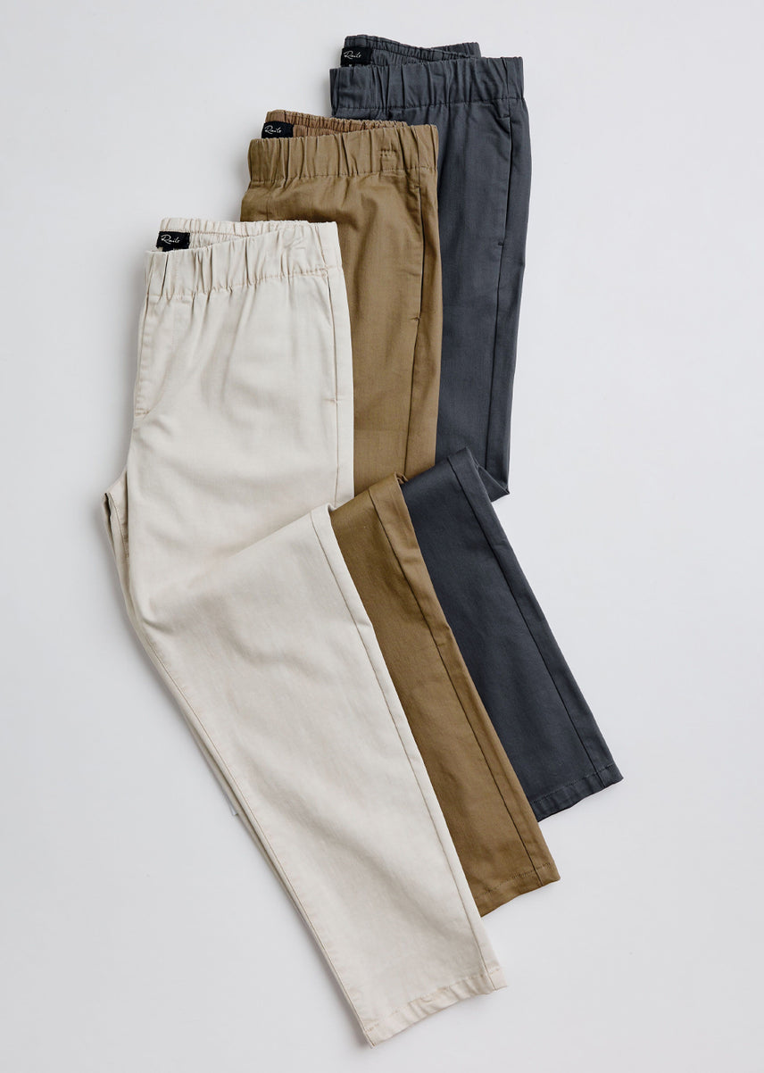 Three pairs of children's elastic-waist pants in cream, tan brown, and dark gray, arranged in a stacked fan layout on a light gray background.