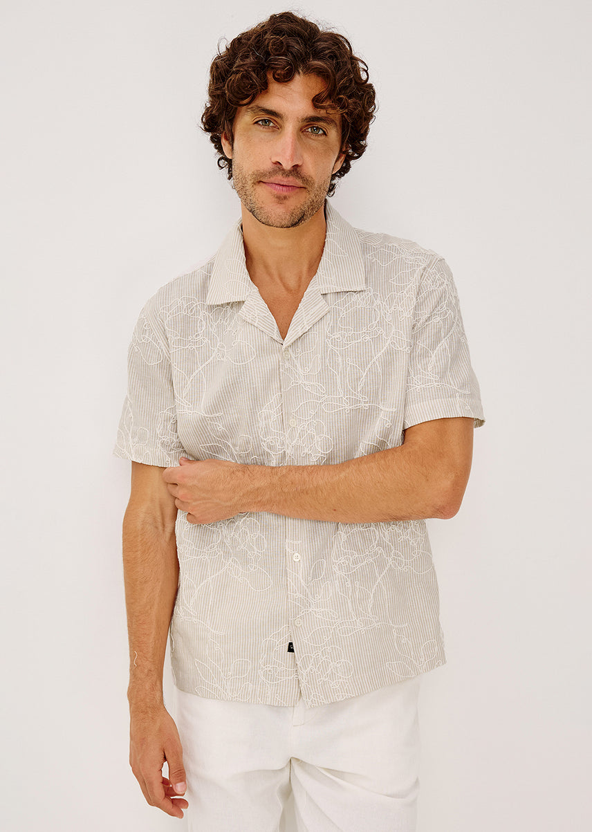 A man wearing a light beige short-sleeve button-up shirt with a white abstract line pattern, paired with white pants, photographed against a plain light background.