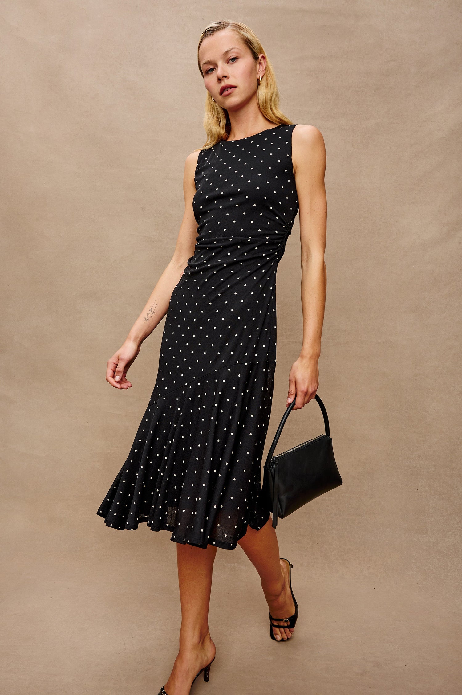 Daija Sleeveless Midi Dress with a Pleated Asymmetrical Hemline, and a Crew Neckline in Polka Dots Colorway - Stylized Front View Featured on Model Holding Purse