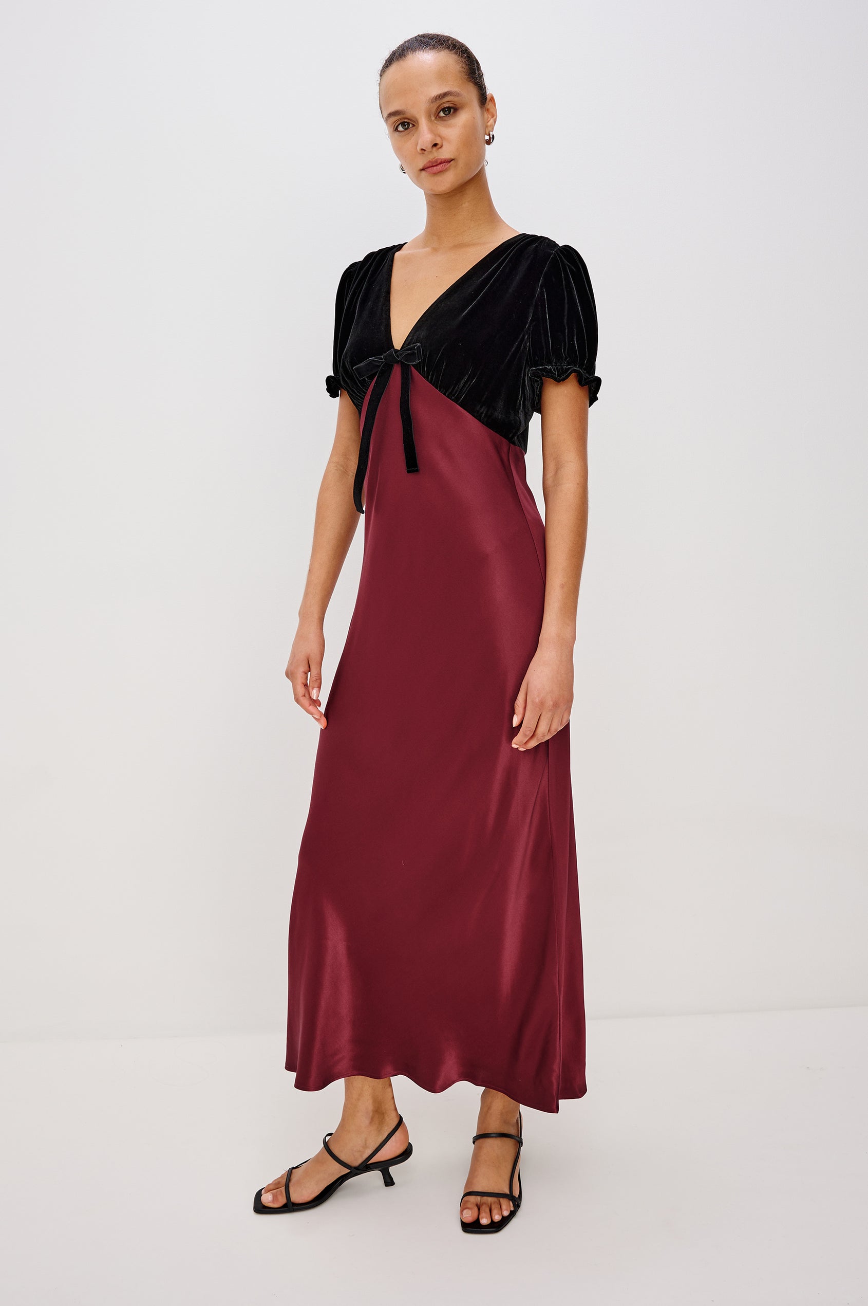 A garnet velvet maxi dress with a fitted V-neck bodice and adjustable center-bust ties. The bias-cut skirt drapes elegantly to ankle length, finished with a back zipper closure. Front View on Model.
