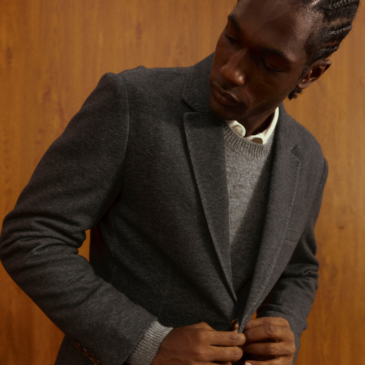 A man wearing a dark gray wool-blend blazer over a gray knit sweater and white collared shirt, standing against a warm wood-paneled background.