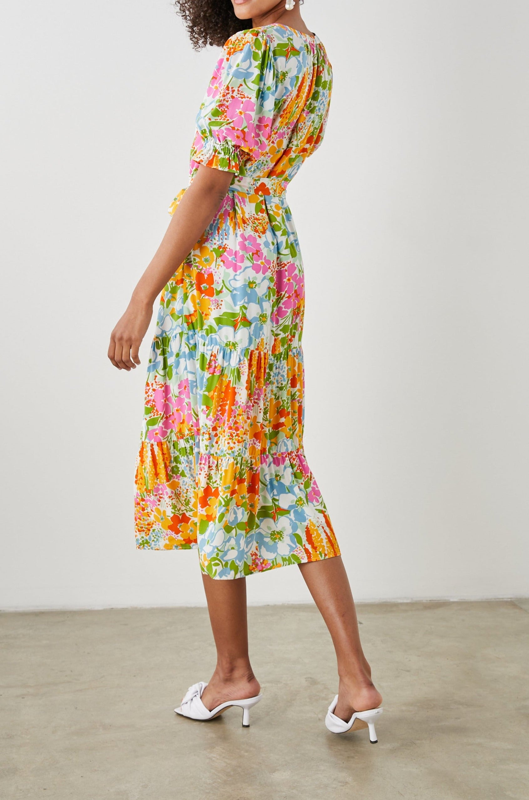 Eliana Midi Dress with Elbow-Length Sleeves with Shirred Cuffs, a V Neck, Buttons Down the Bodice, a Fabric Tie at the Waist  and a Tiered Skirt in Day Garden Colorway, a Floral Pattern in Blue, Pink and Oranges on White - Back Full Body View Featured on Model