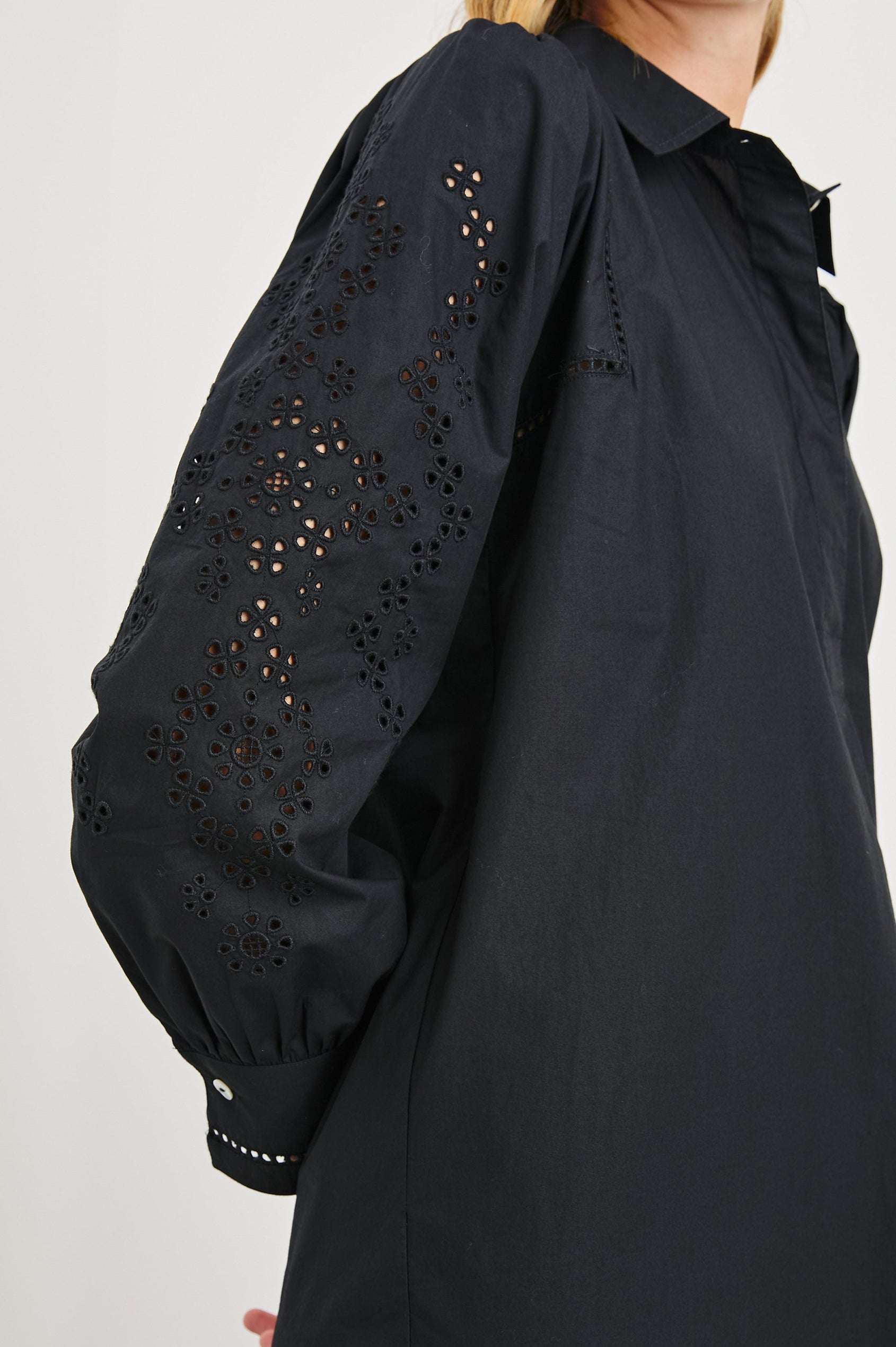 Fernanda Long-Sleeve Mini Dress with Button Cuffs, a Collar, a Placket with Buttons, a Scalloped Hem, a Floral Eyelet Design on the Sleeves and a Floral and Geometric Eyelet Design on the Hem in Black Eyelet Colorway - Side View Featured on Model