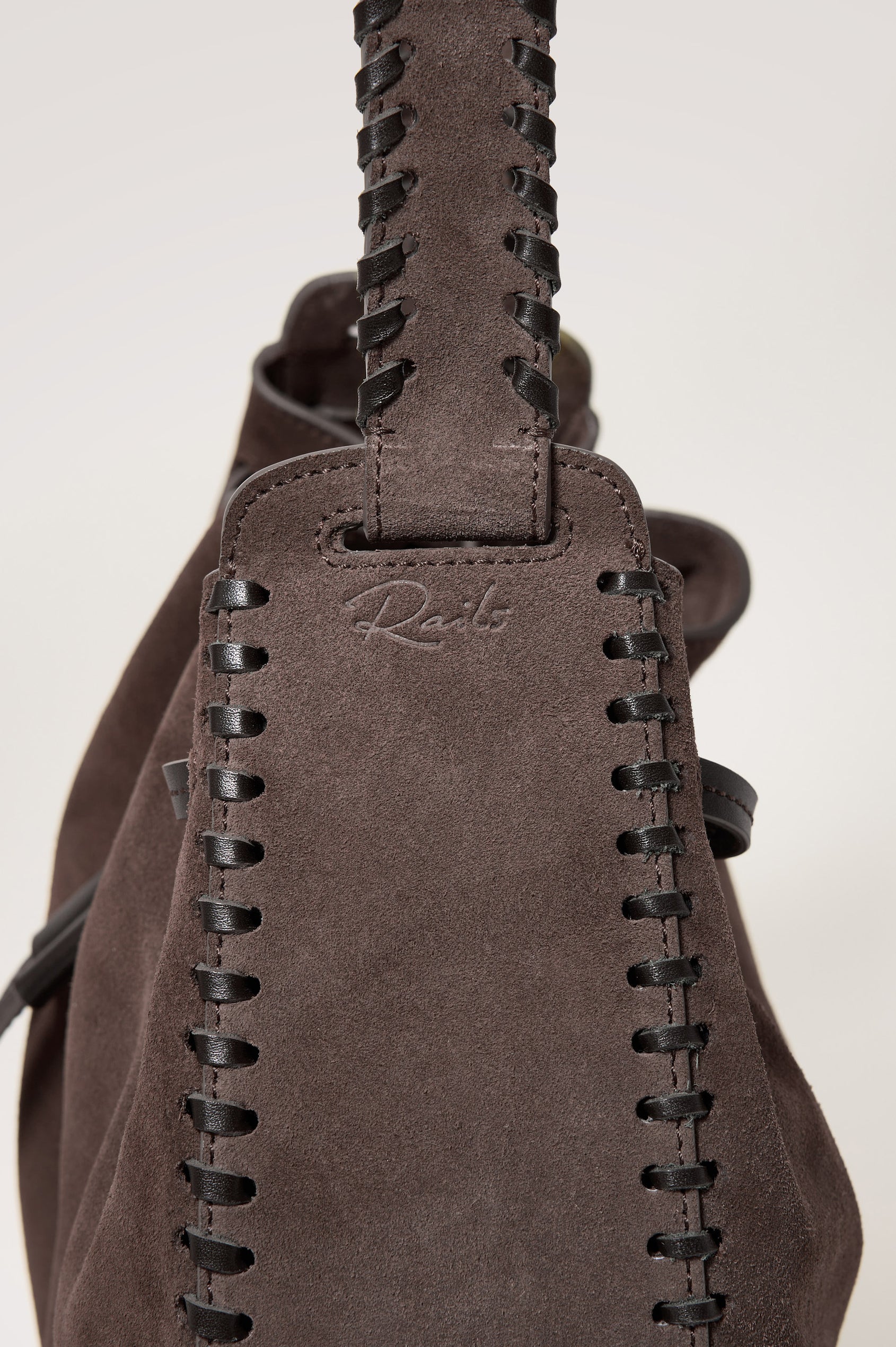 Firenze Bucket Bag in Suede With Whipstitch Trim, Drawcord Closure, and Interior Zip Pocket in Dark Brown Suede Colorway - close up side view of exterior, showing rails logo at the base of the strap.