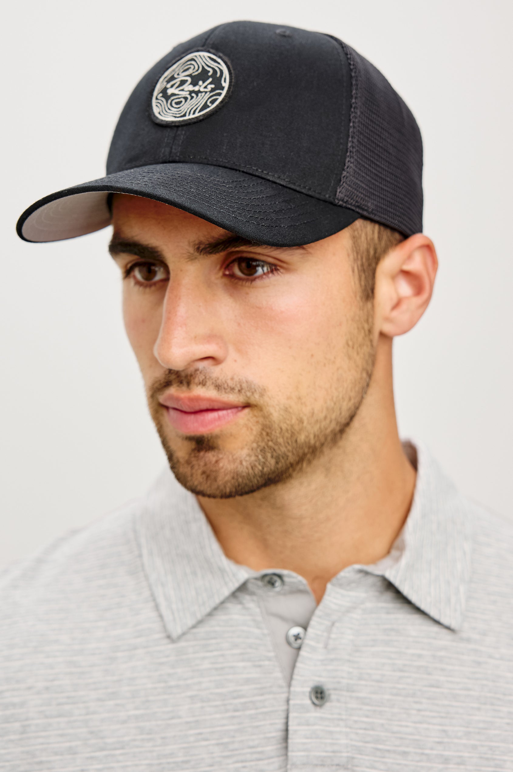 Golf Hat With Embroidered Branded Patch at Front, Adjustable Snapback, Mesh Back, and 6-Panel Construction in Black Colorway - Upper Body Front View Featured on Model