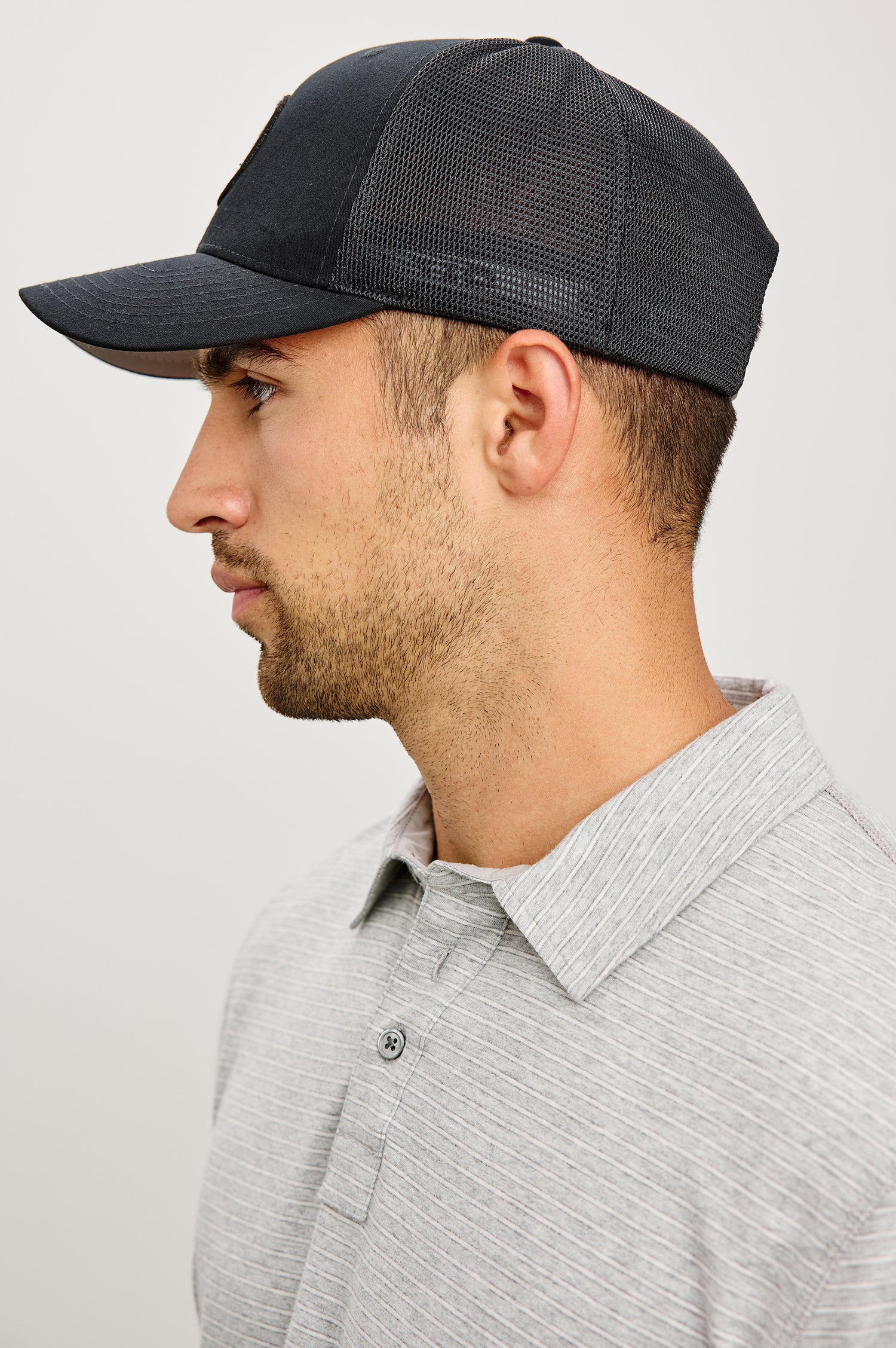 Golf Hat With Embroidered Branded Patch at Front, Adjustable Snapback, Mesh Back, and 6-Panel Construction in Black Colorway - Upper Body Side View Featured on Model