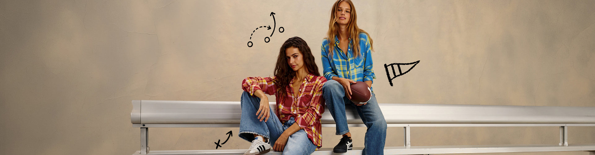 Two women sitting on a silver metal bleacher bench against a textured beige wall. The woman on the left wears a red and brown plaid shirt with blue jeans, and the woman on the right wears a blue and yellow plaid shirt with blue jeans.