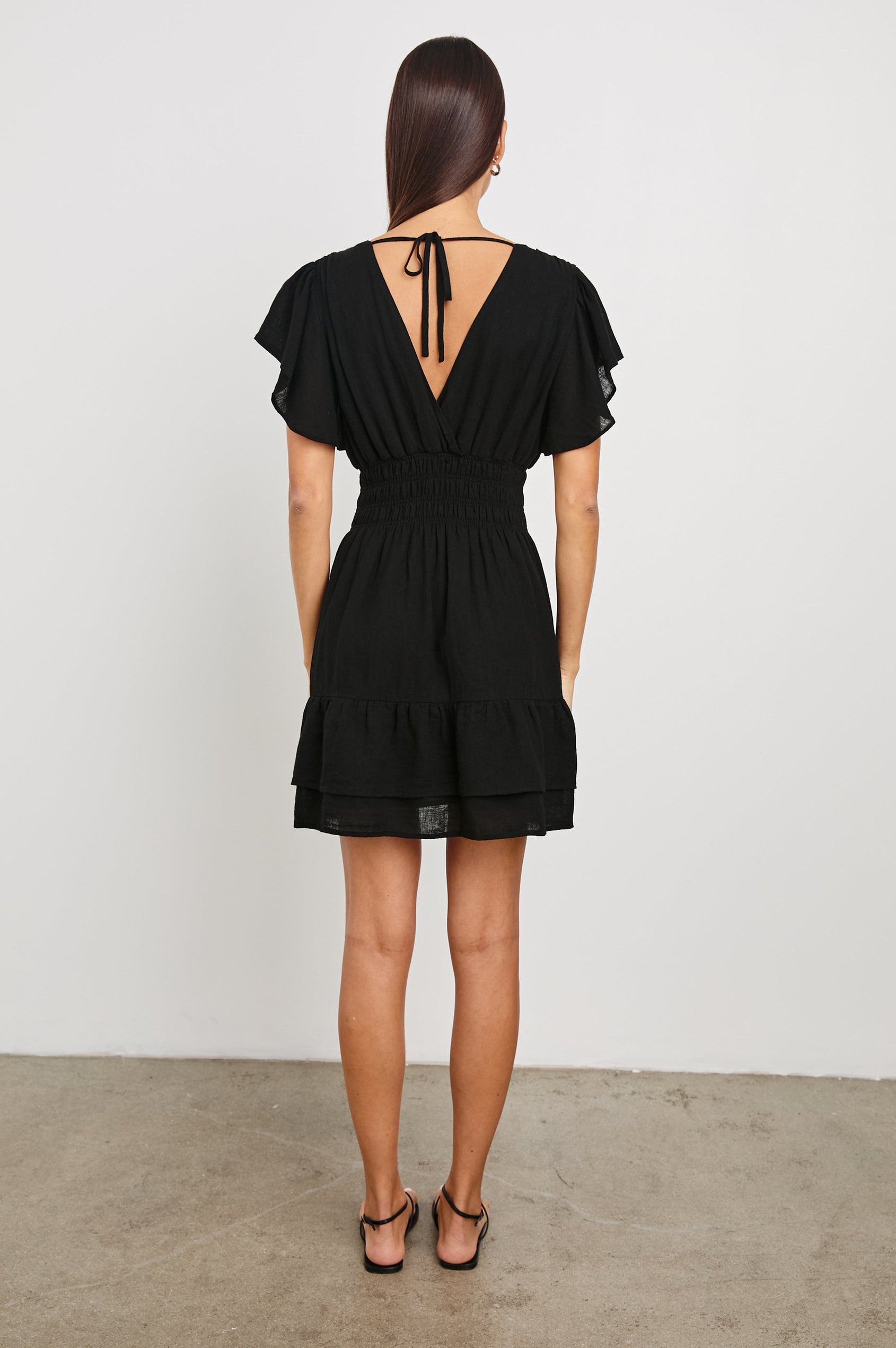 Kimora Above-Knee Dress with Flutter Sleeves, a V Neckline, a Cinched Waist and a Tiered Skirt in Black Colorway - Back Full Body View Featured on Model