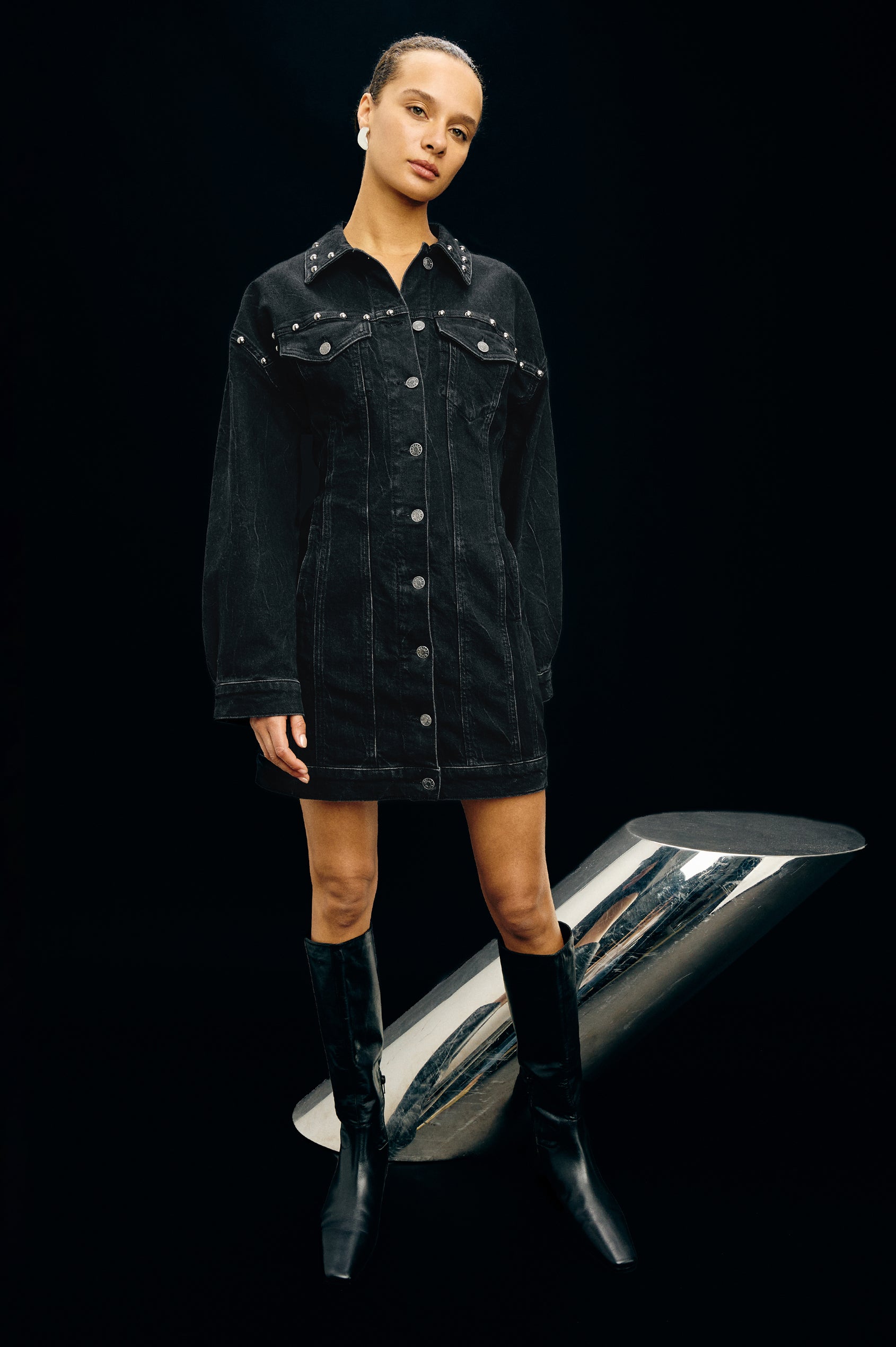 Ladera is a trucker style denim mini dress with long sleeves, drop shoulders, relaxed balloon sleeves, nail-back studs, and front button closures, with a subtle A-line shape and adjustable back strap in Black Stone Studded colorway. Front full body view featured on model.