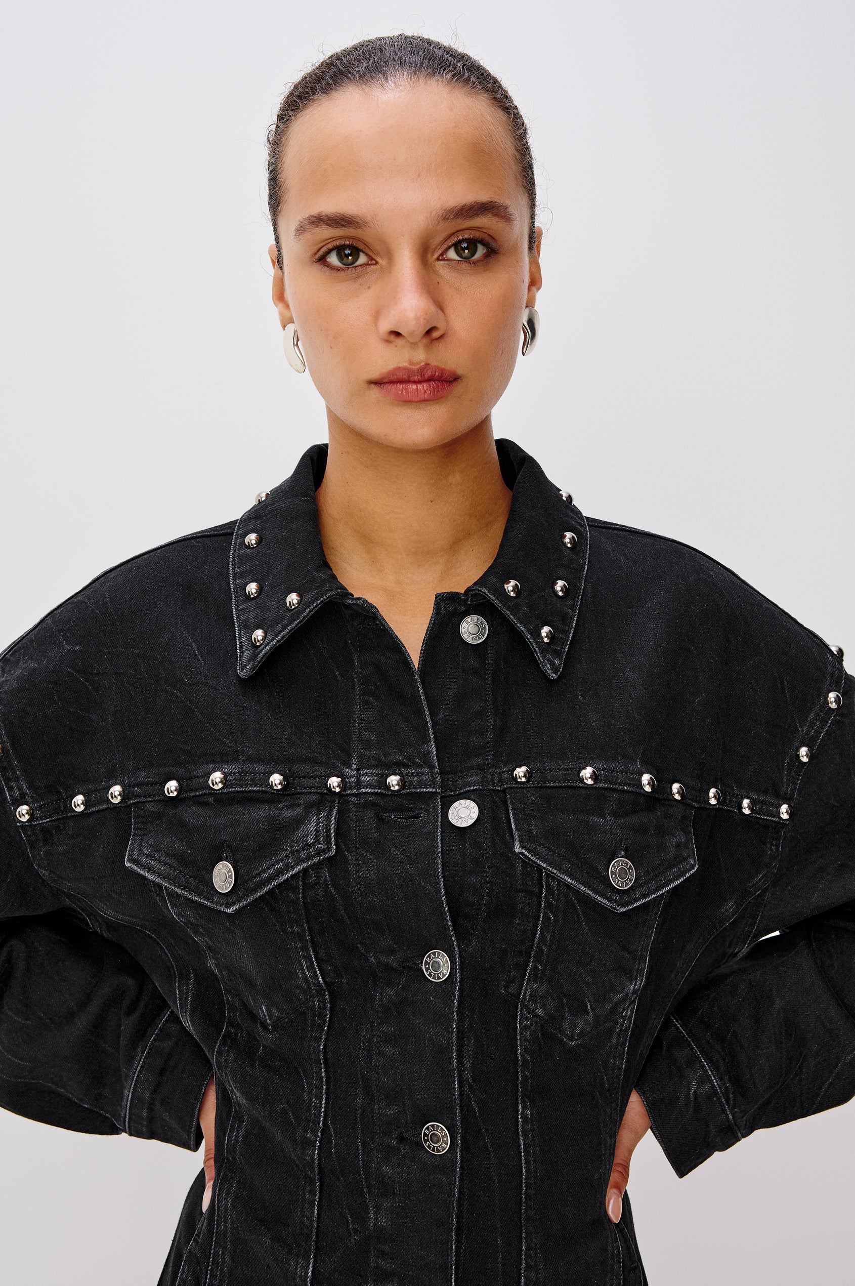 Ladera is a trucker style denim mini dress with long sleeves, drop shoulders, relaxed balloon sleeves, nail-back studs, and front button closures, with a subtle A-line shape and adjustable back strap in Black Stone Studded colorway. Front detail view featured on model.