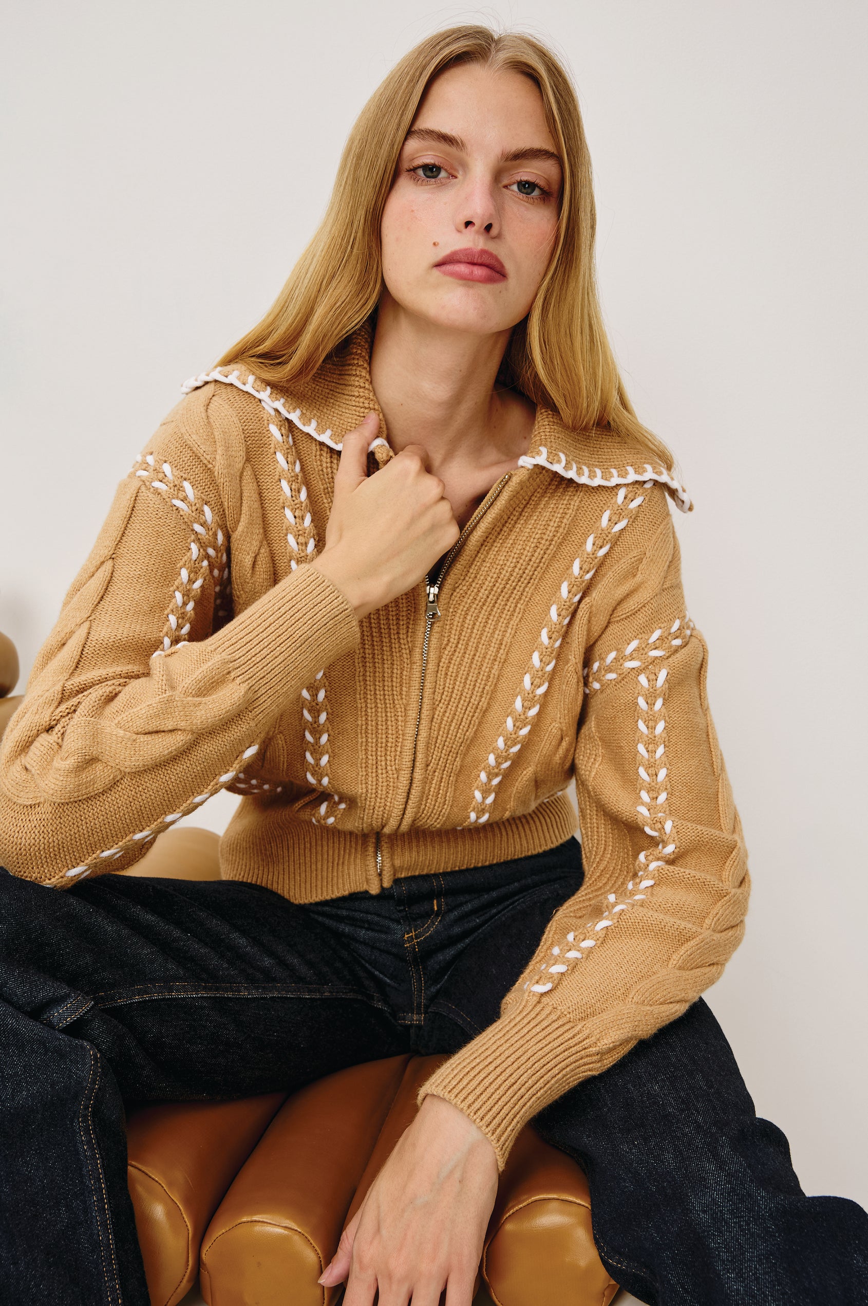 Lune Sweater in Cable Knit With Contrast Blanket Stitch Detailing, Stand Collar, Front Zipper, and Long Sleeves in Camel Ivory Blanket Stitch Colorway - Upper Body Front View Featured on Model Sitting