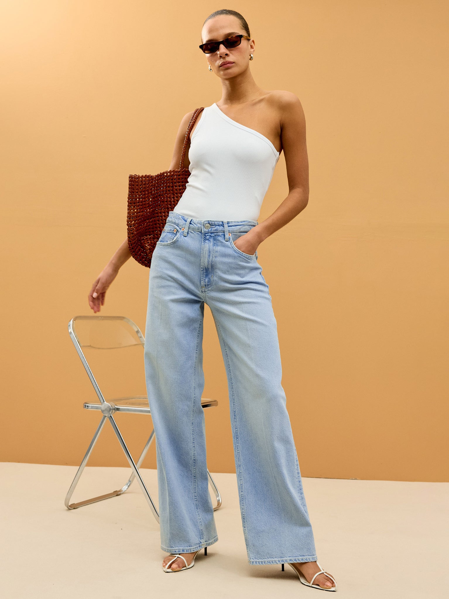 Woman wearing a white one-shoulder top and light blue jeans against a beige background