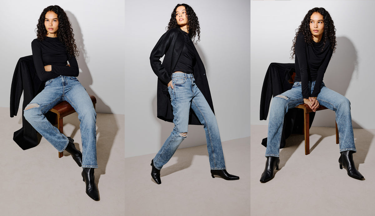 A model wearing a black longline coat, black top, light-wash distressed blue jeans, and black ankle boots, posing in a studio setting.