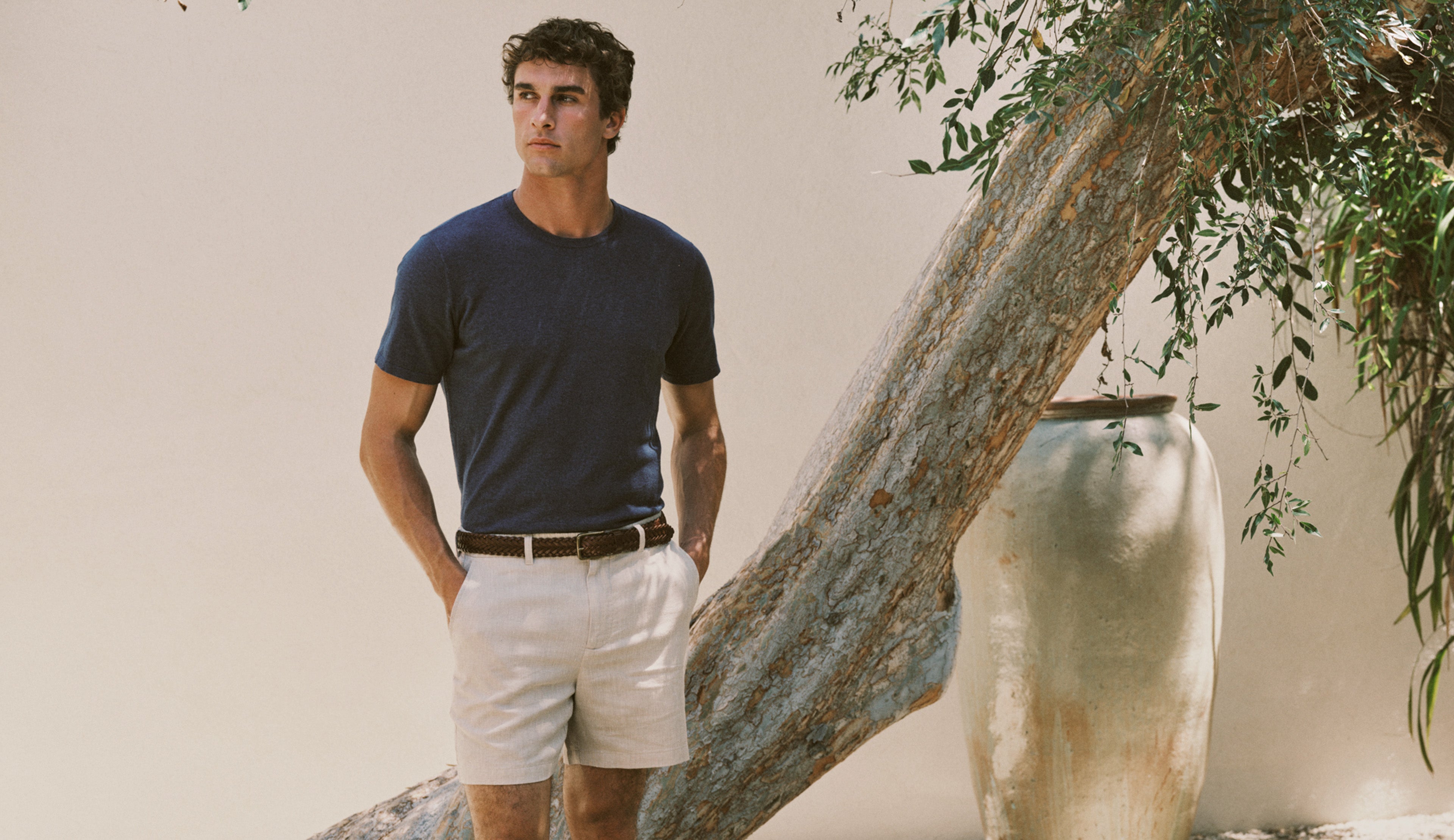 A man wearing a dark navy blue crewneck t-shirt and off-white shorts, standing outdoors next to a large tree trunk and a decorative ceramic pot.
