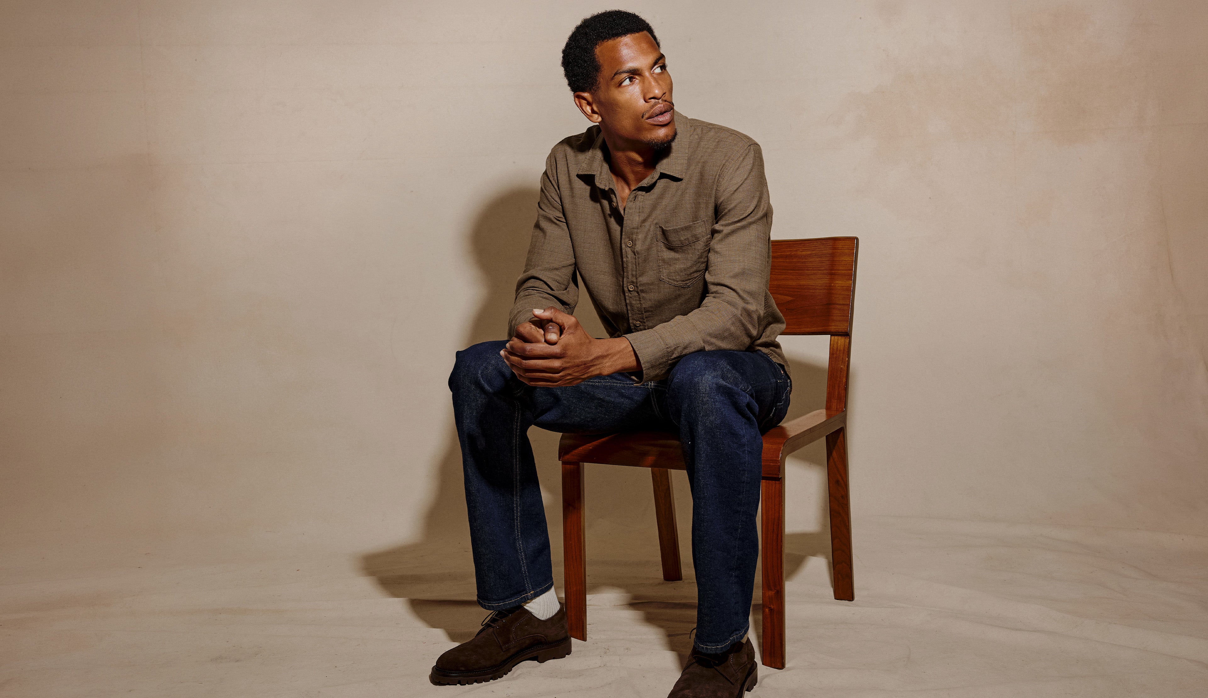 A man sits on a wooden chair against a textured beige studio backdrop, wearing a brown button-up shirt, dark blue jeans, and brown shoes.