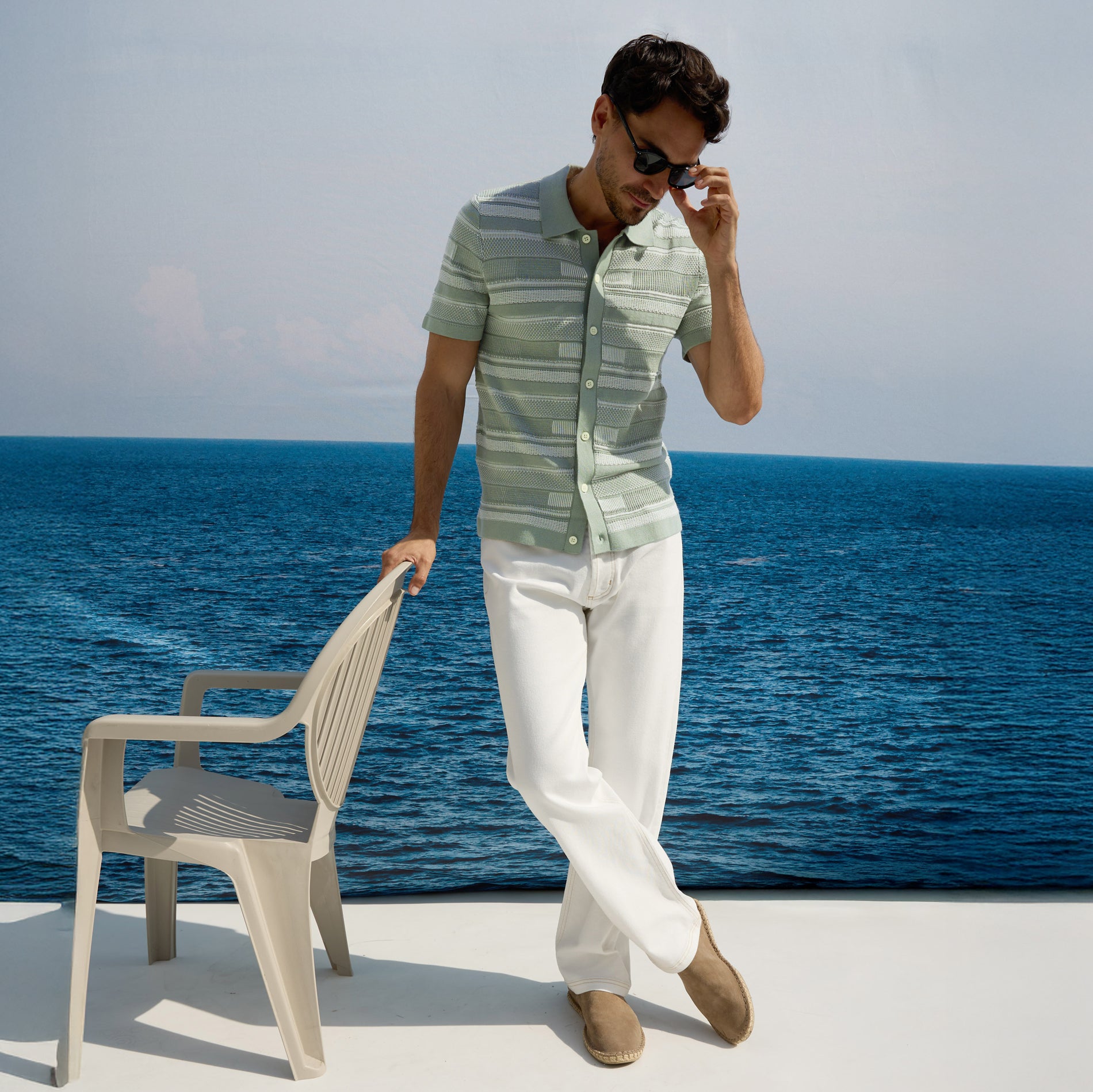 Man in striped shirt and white pants standing by the ocean with a chair.