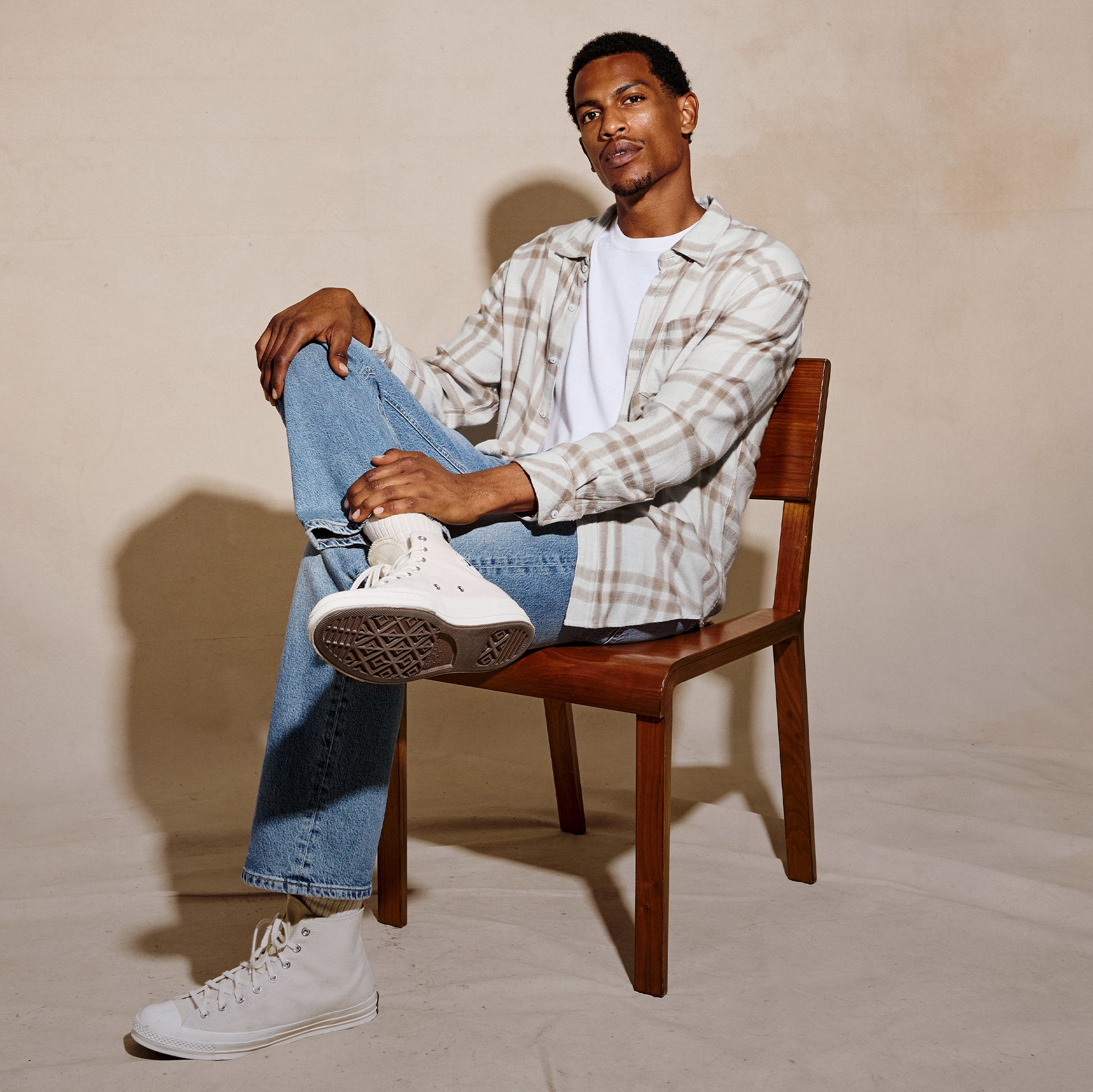A man sits on a wooden chair wearing a beige and white plaid flannel shirt over a white t-shirt, blue jeans, and white high-top sneakers against a warm beige studio backdrop.