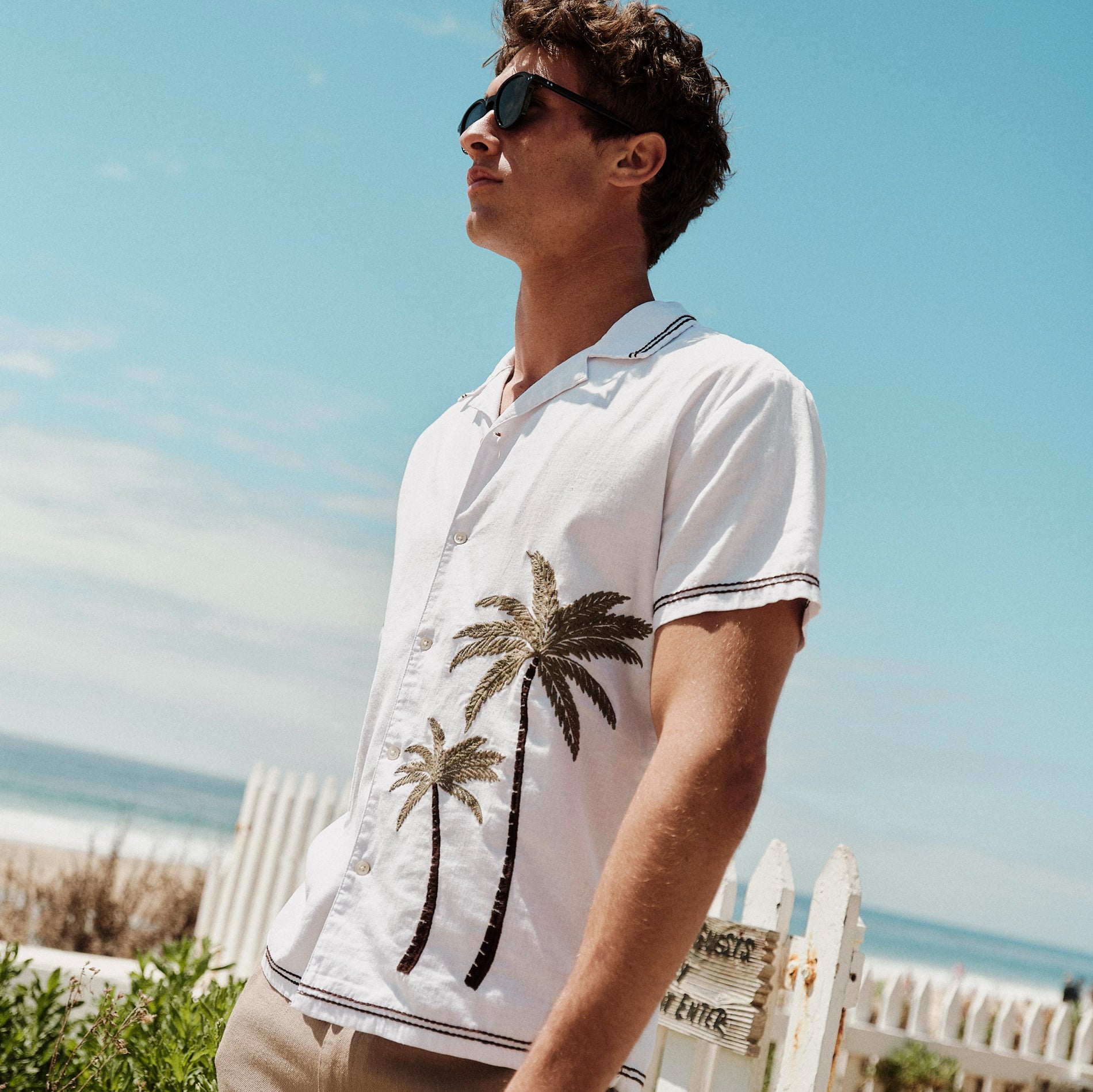 Man wearing a white polo shirt with palm tree design on a beach.