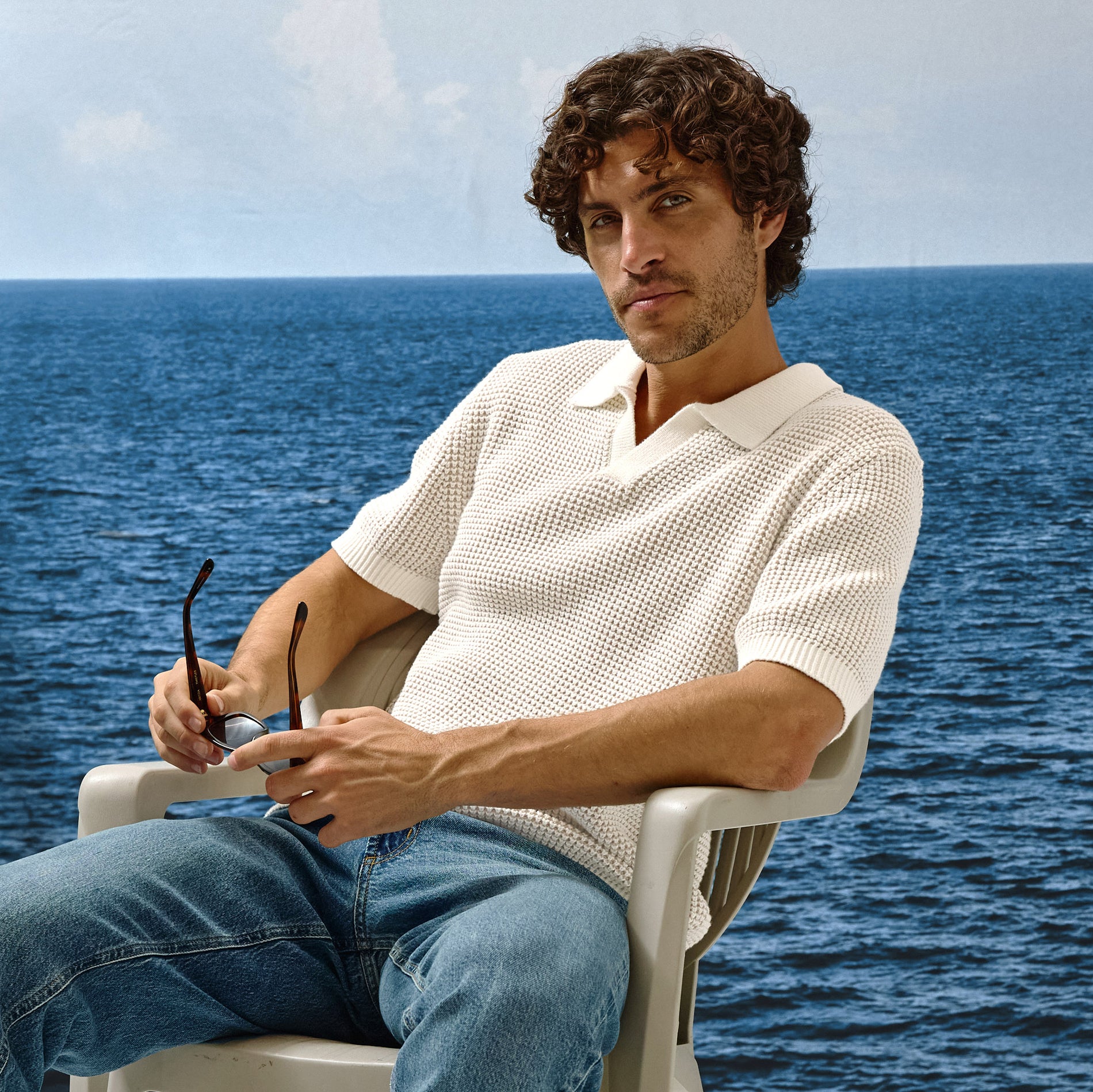Man sitting on a chair by the ocean wearing a white shirt and blue jeans.