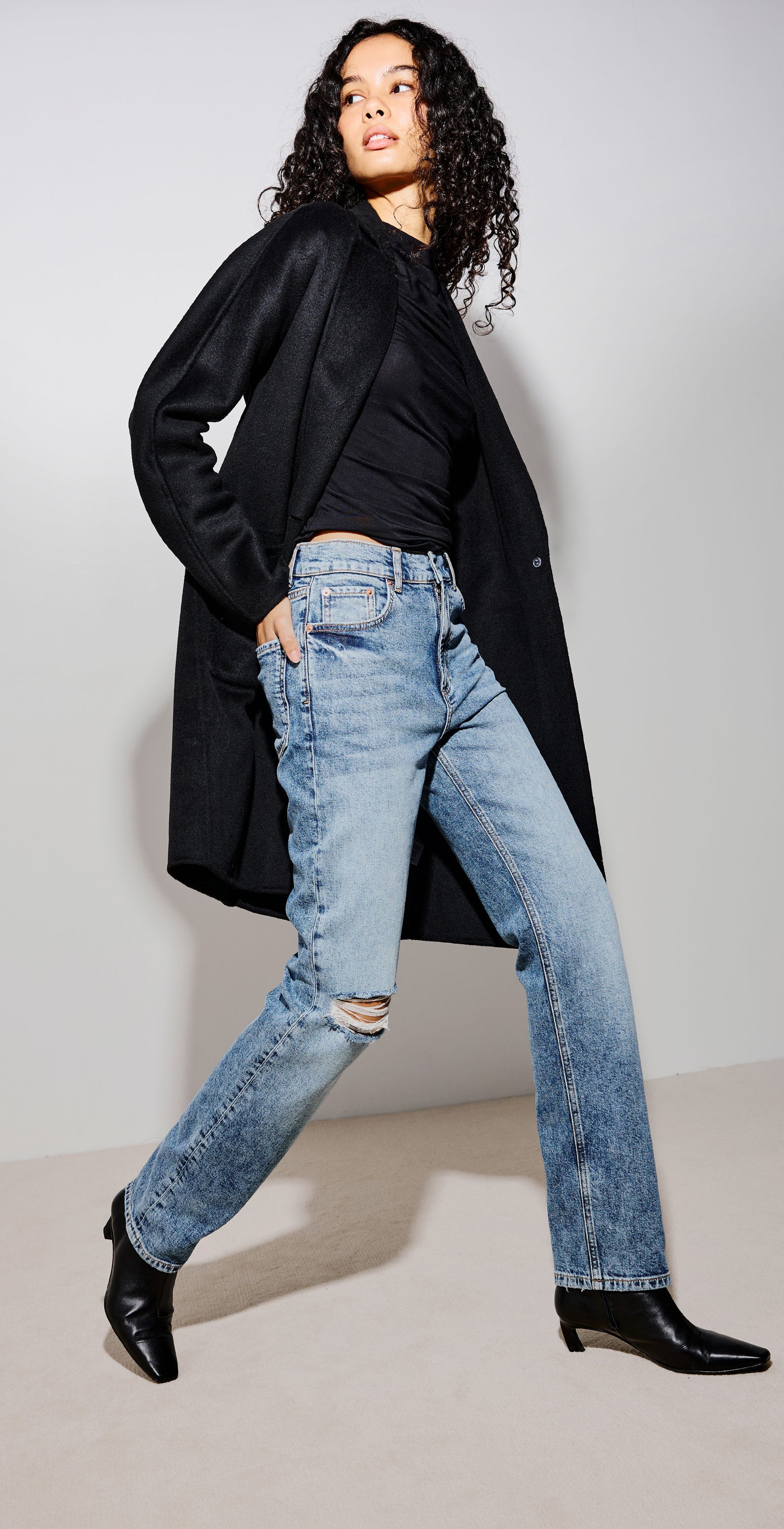 A model wearing a black longline coat, black top, light-wash distressed blue jeans, and black ankle boots, posing in a studio setting.