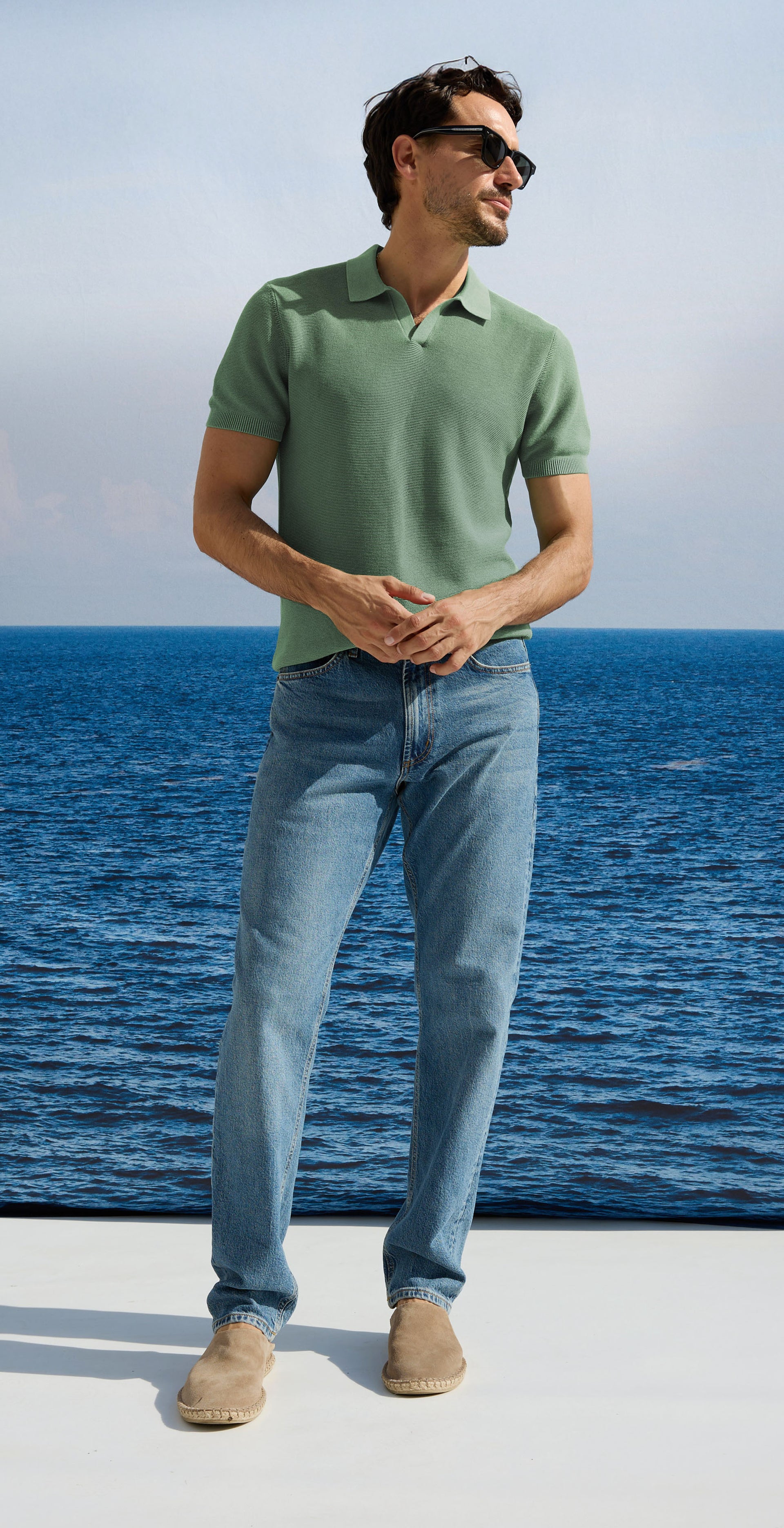 Man wearing a green polo shirt and blue jeans standing by the ocean.