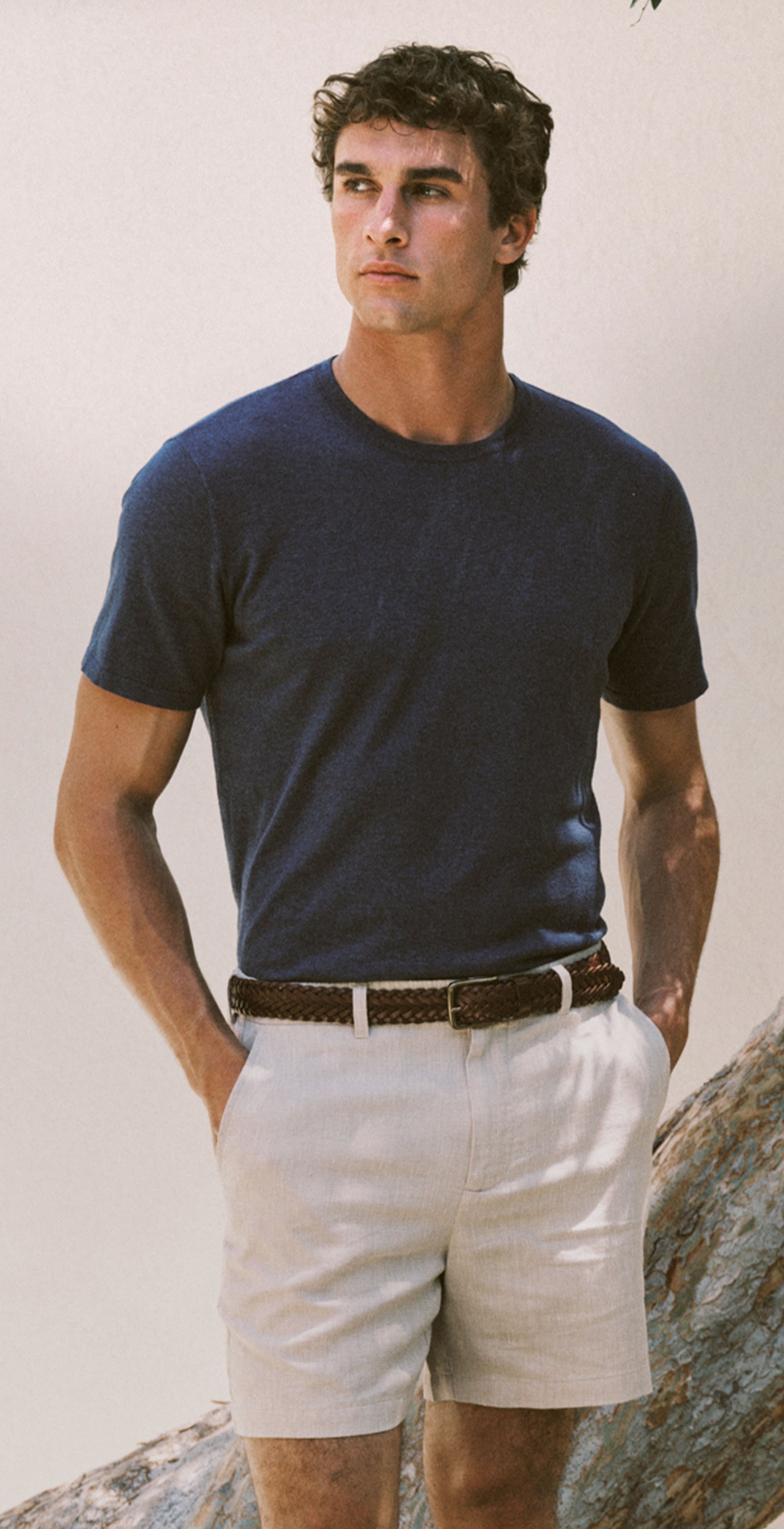 A man wearing a dark navy blue crewneck t-shirt and off-white shorts, standing outdoors next to a large tree trunk and a decorative ceramic pot.