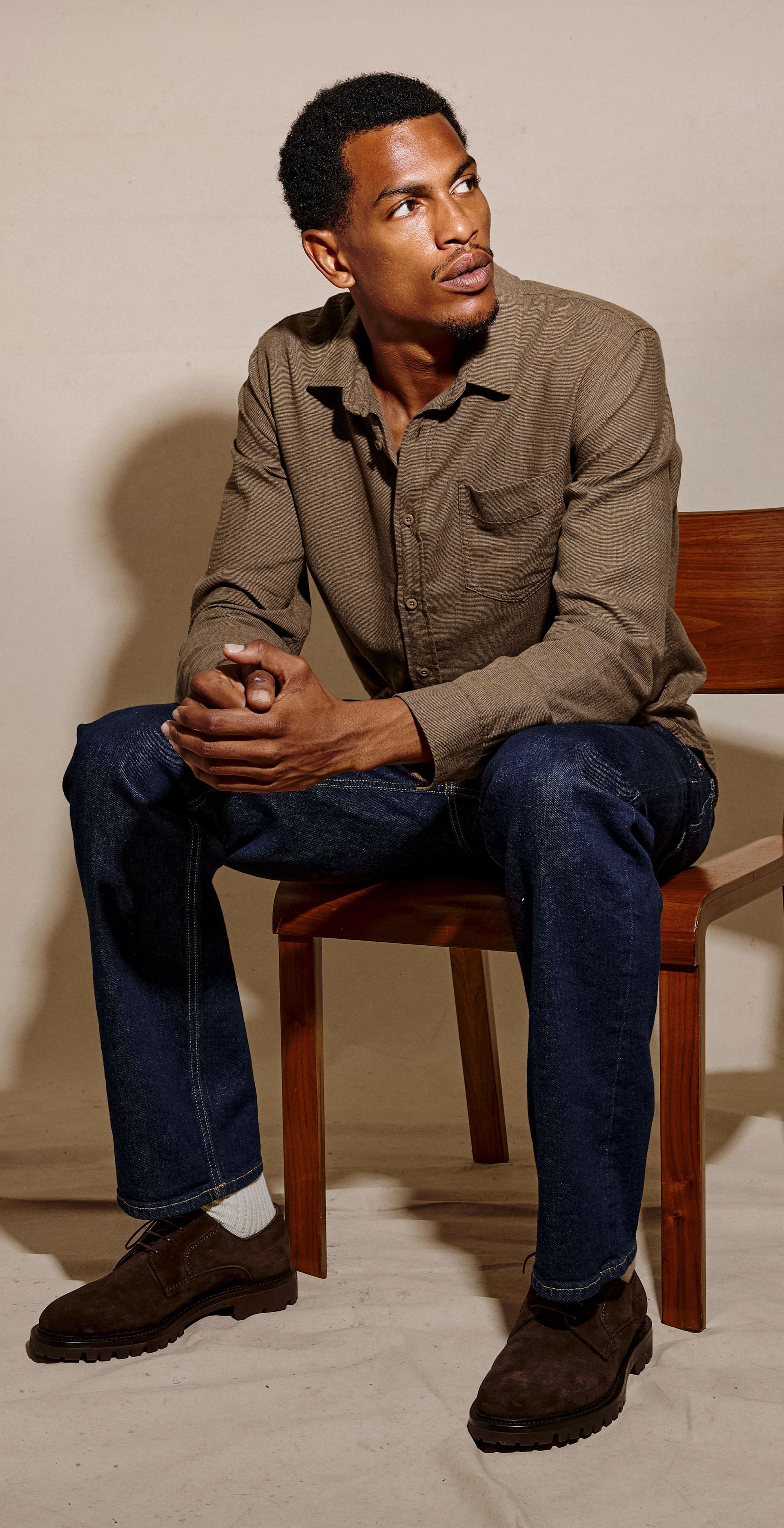A man sits on a wooden chair against a textured beige studio backdrop, wearing a brown button-up shirt, dark blue jeans, and brown shoes.